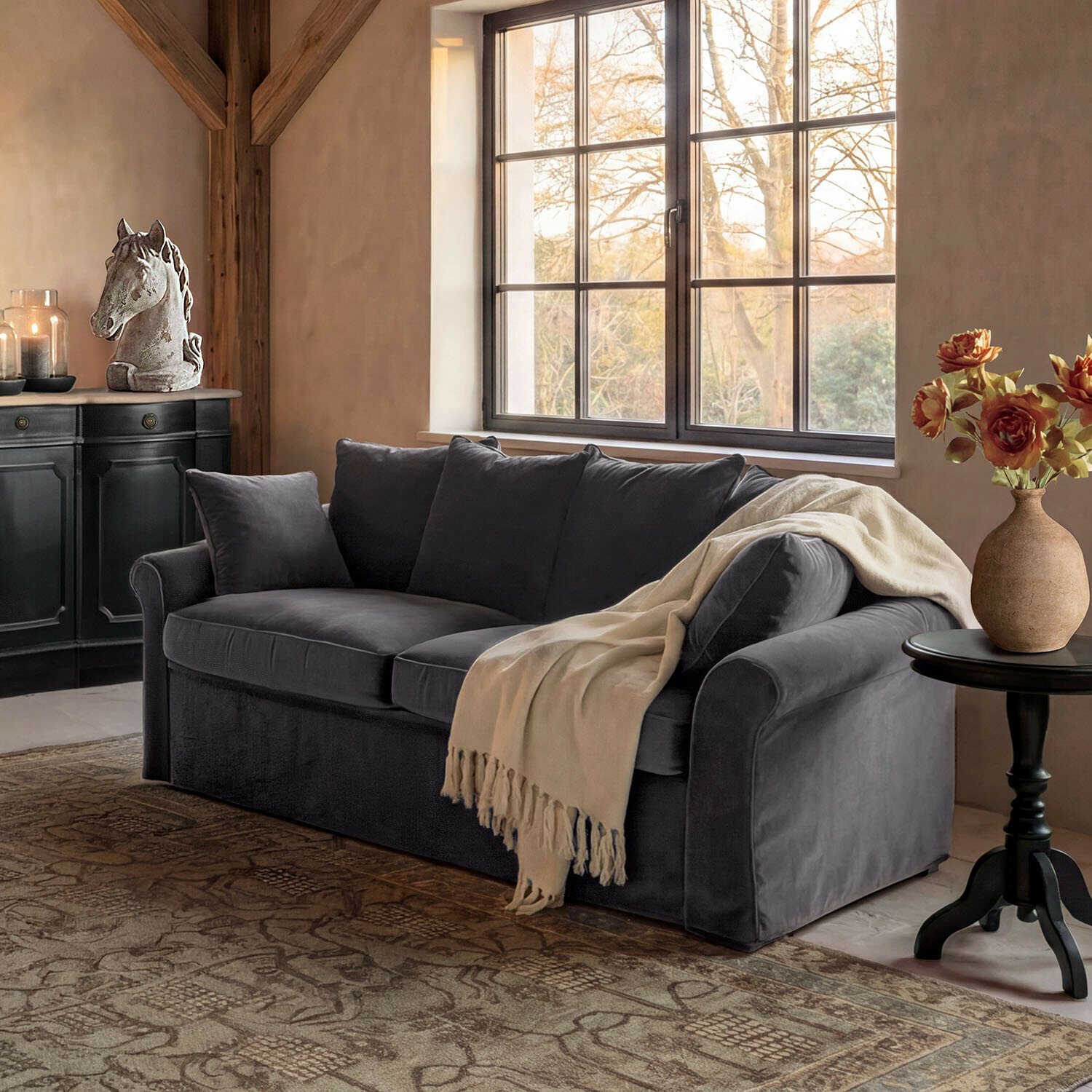 Sofa Long Beach Graues Samtsofa mit cremefarbenem Plaid in gemütlichem Wohnzimmer mit Pferdekopf-Dekor und Blumen