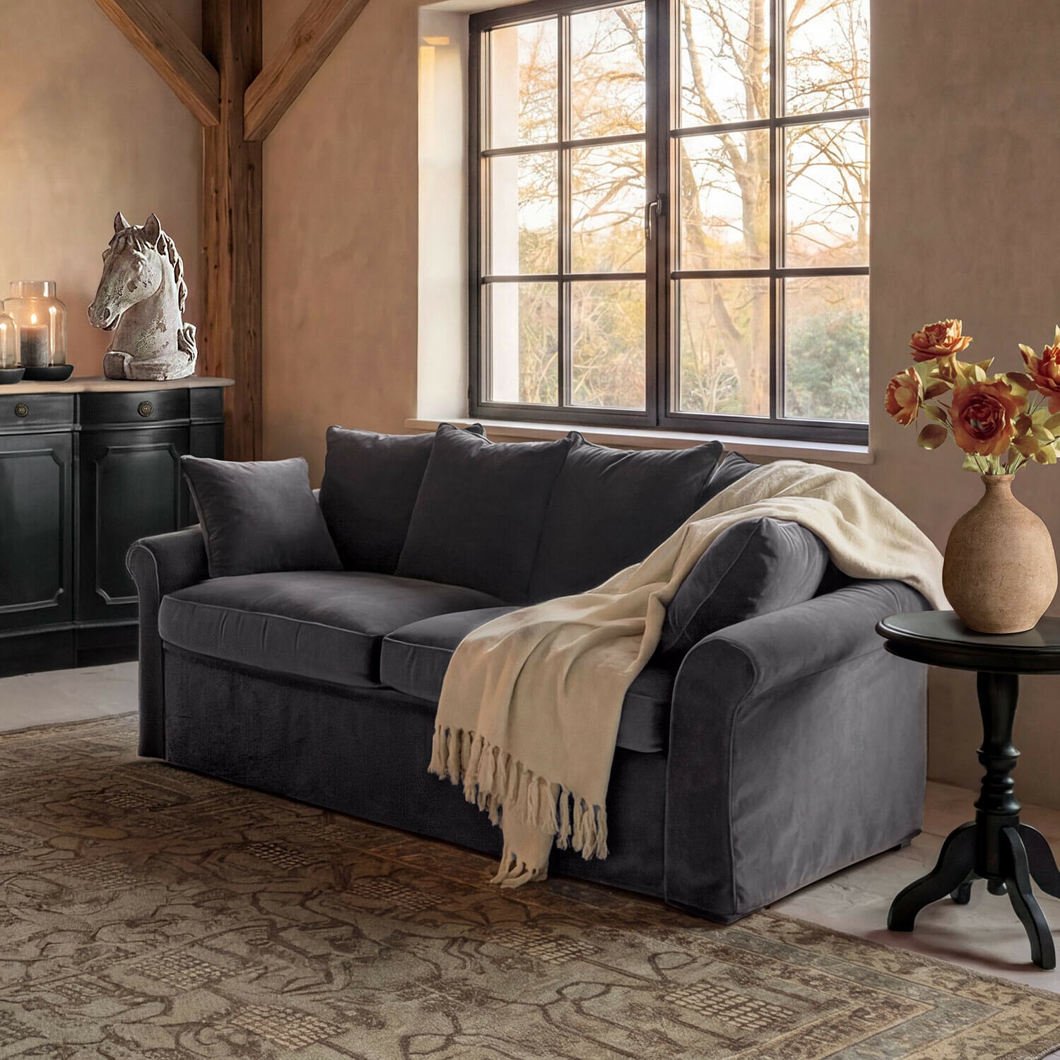 Sofa Long Beach Graues Samtsofa mit cremefarbenem Plaid in gemütlichem Wohnzimmer mit Pferdekopf-Dekor und Blumen