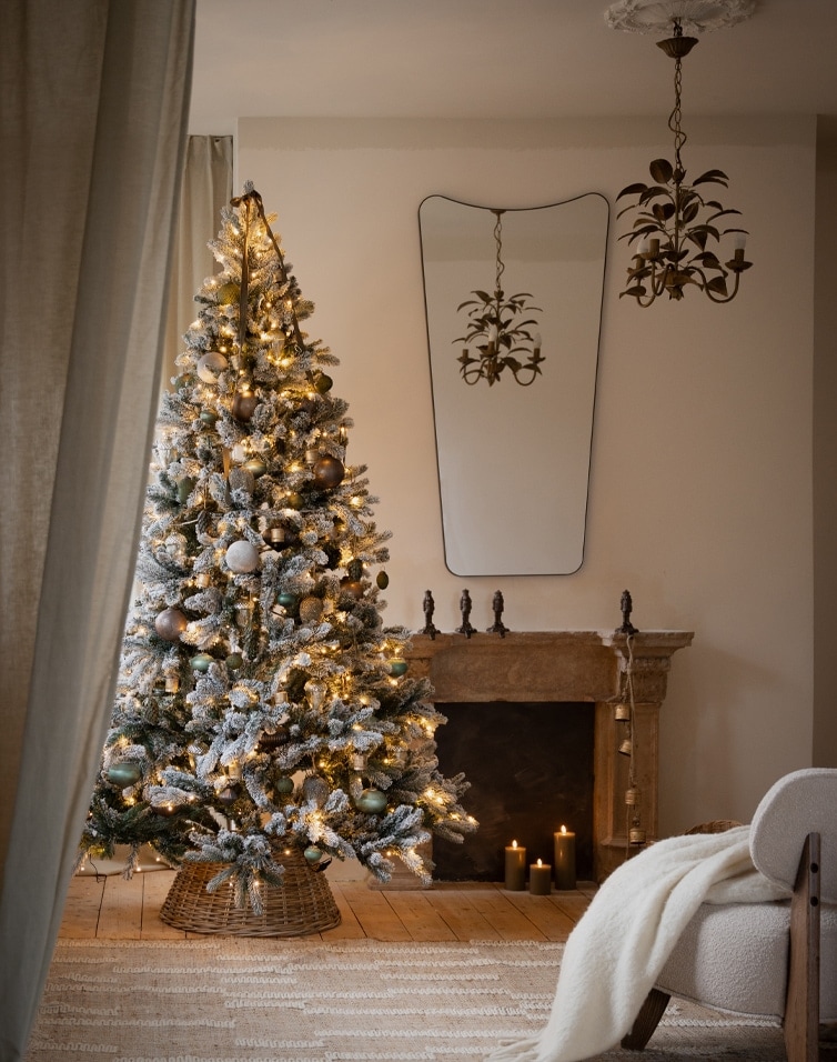 Oberflockierter Weihnachtsbaum mit Lichterkette und Kugeln im Korb vor gemütlichem Kaminzimmer