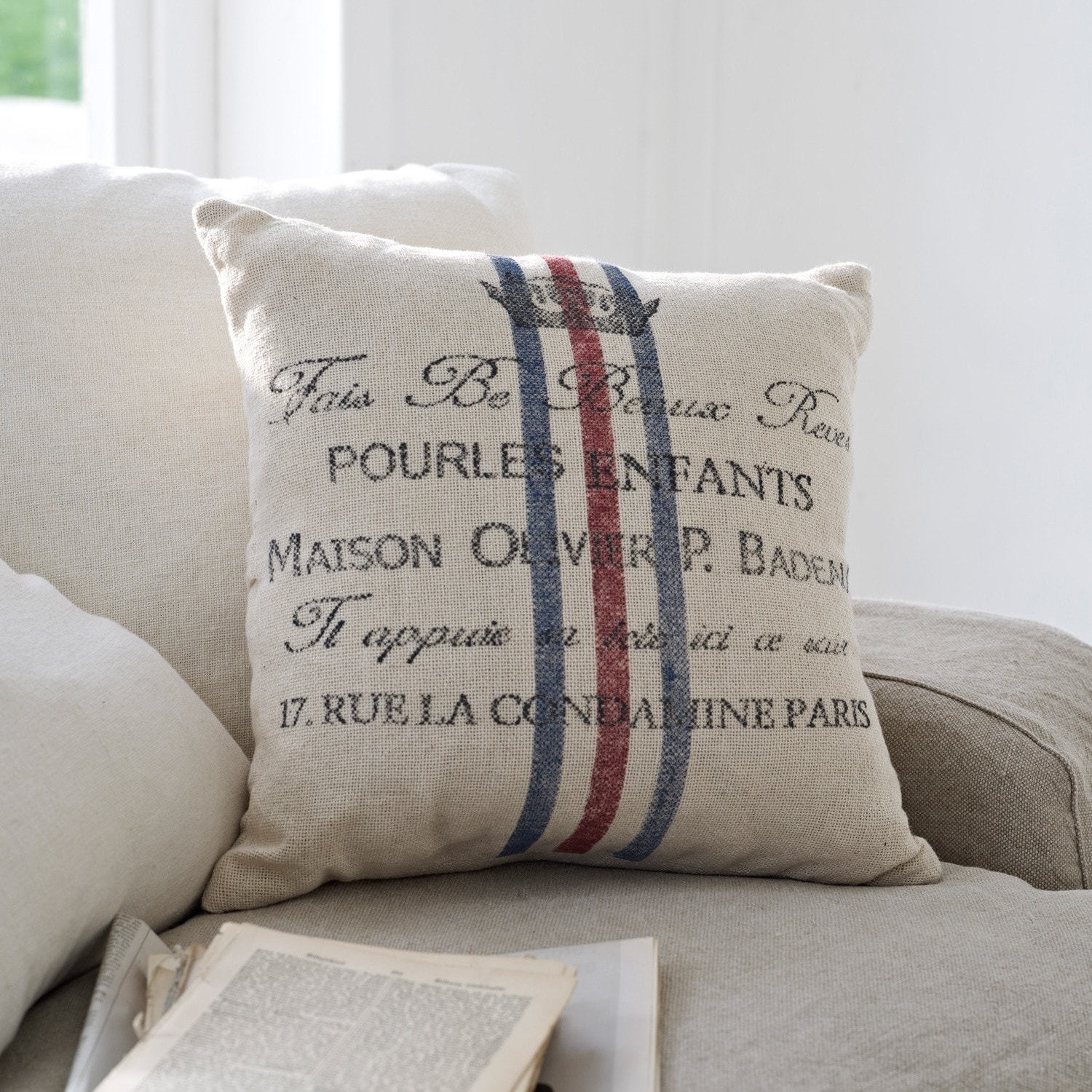 Kissen Pourl Beige Dekokissen aus Stoff mit Schrift und rot-blauen Streifen auf hellem Sofa im Wohnzimmer