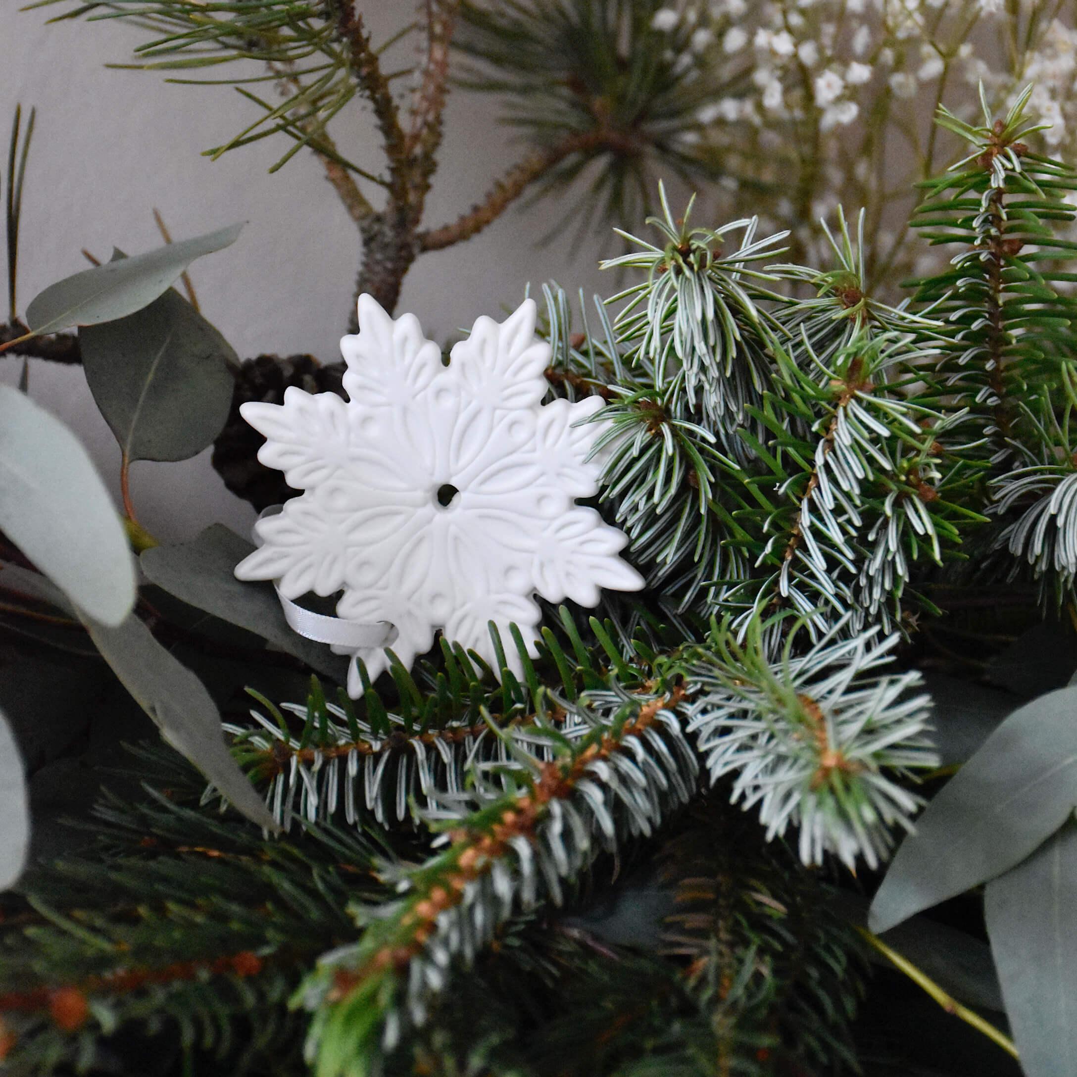 Weiße Keramik-Weihnachtsdekoration in Schneeflockenform auf grünen Tannenzweigen