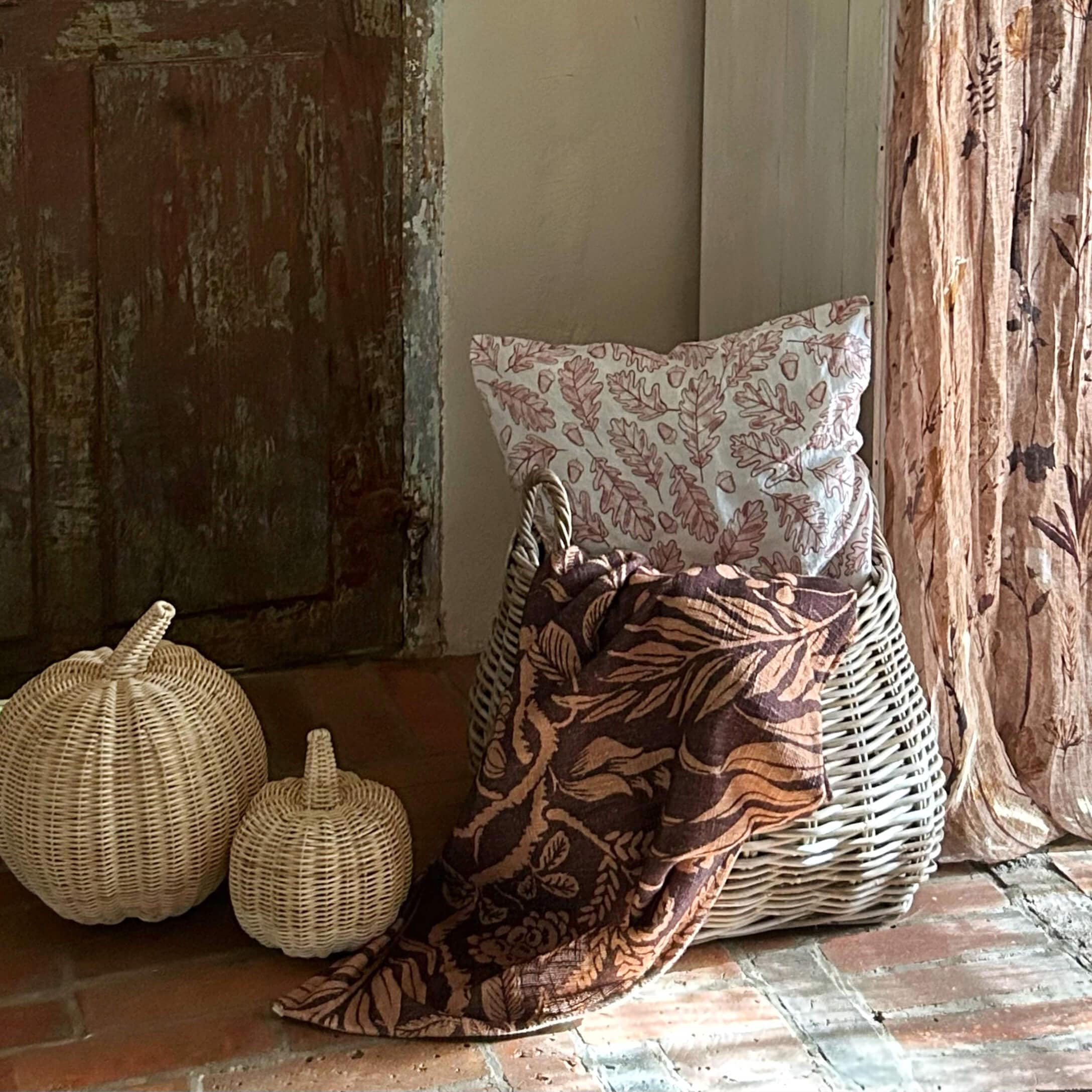 Weißer Korb aus Weide mit Blattmuster-Kissen und braunem Deckenstoff auf Ziegelboden vor altem Holztür