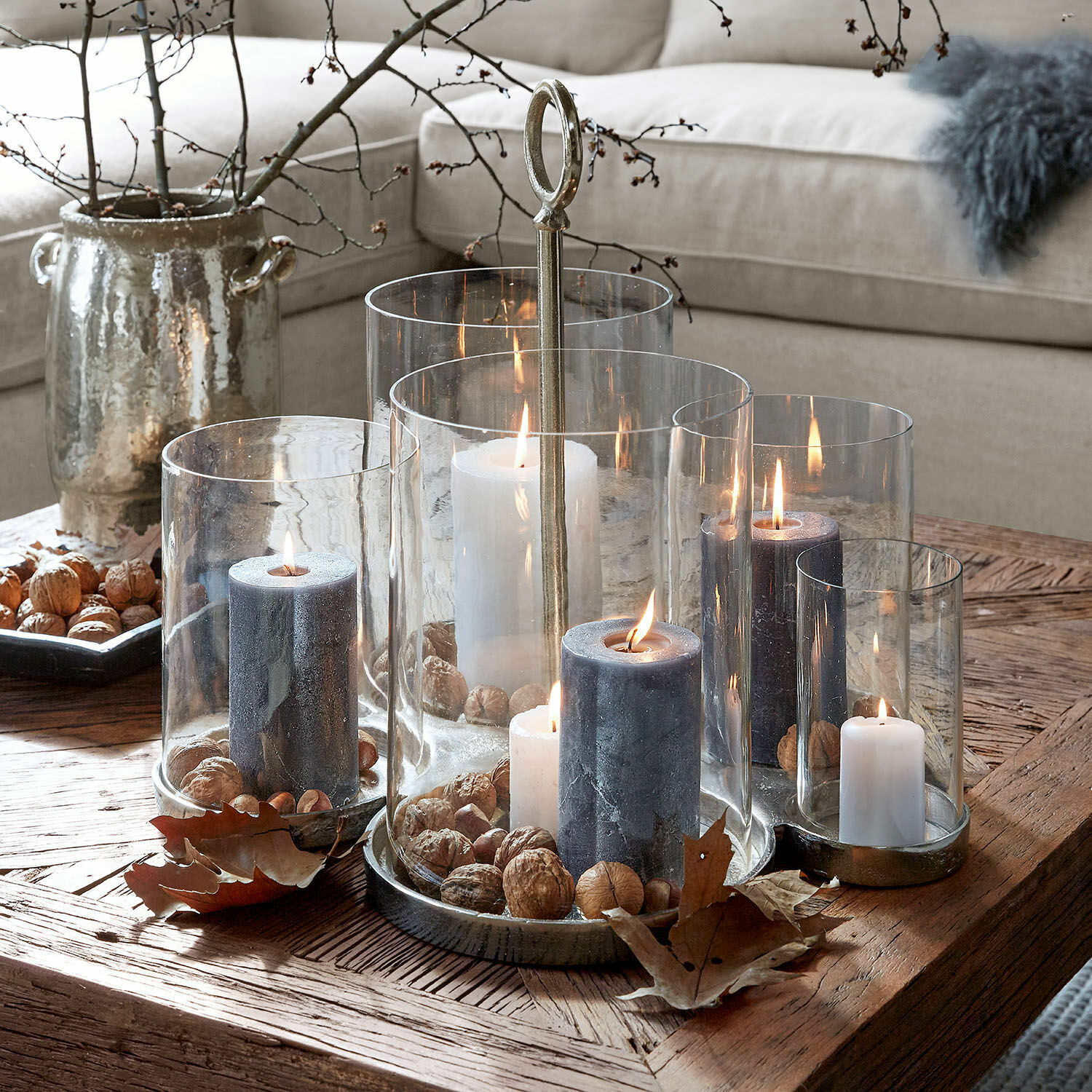 Windlicht Gjaloran Set aus Glas- und Metall-Kerzenhaltern mit weißen und grauen Kerzen und Nüssen auf Holztisch