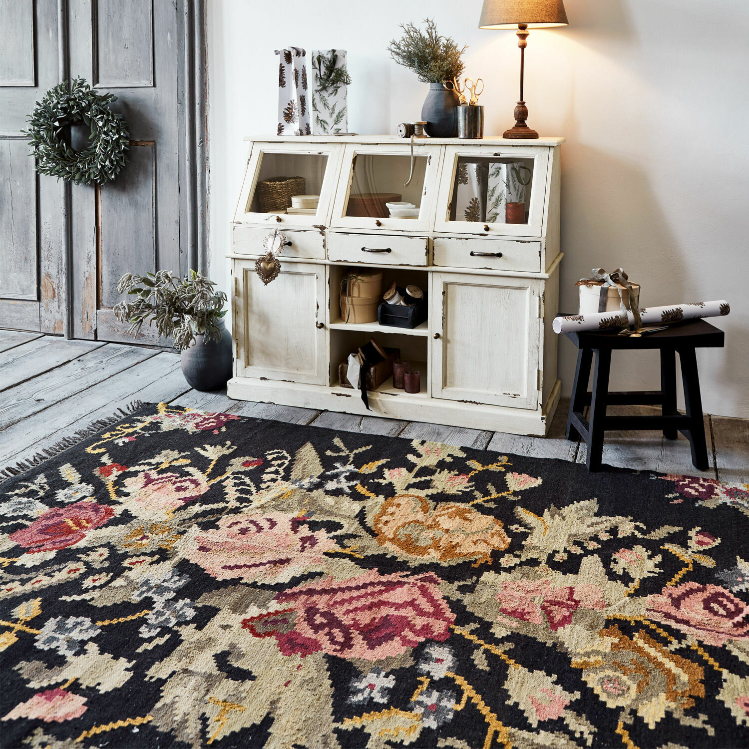 Dunkler Teppich mit floralem Muster vor weißem Vintage-Holzschrank und dekorativer Lampe