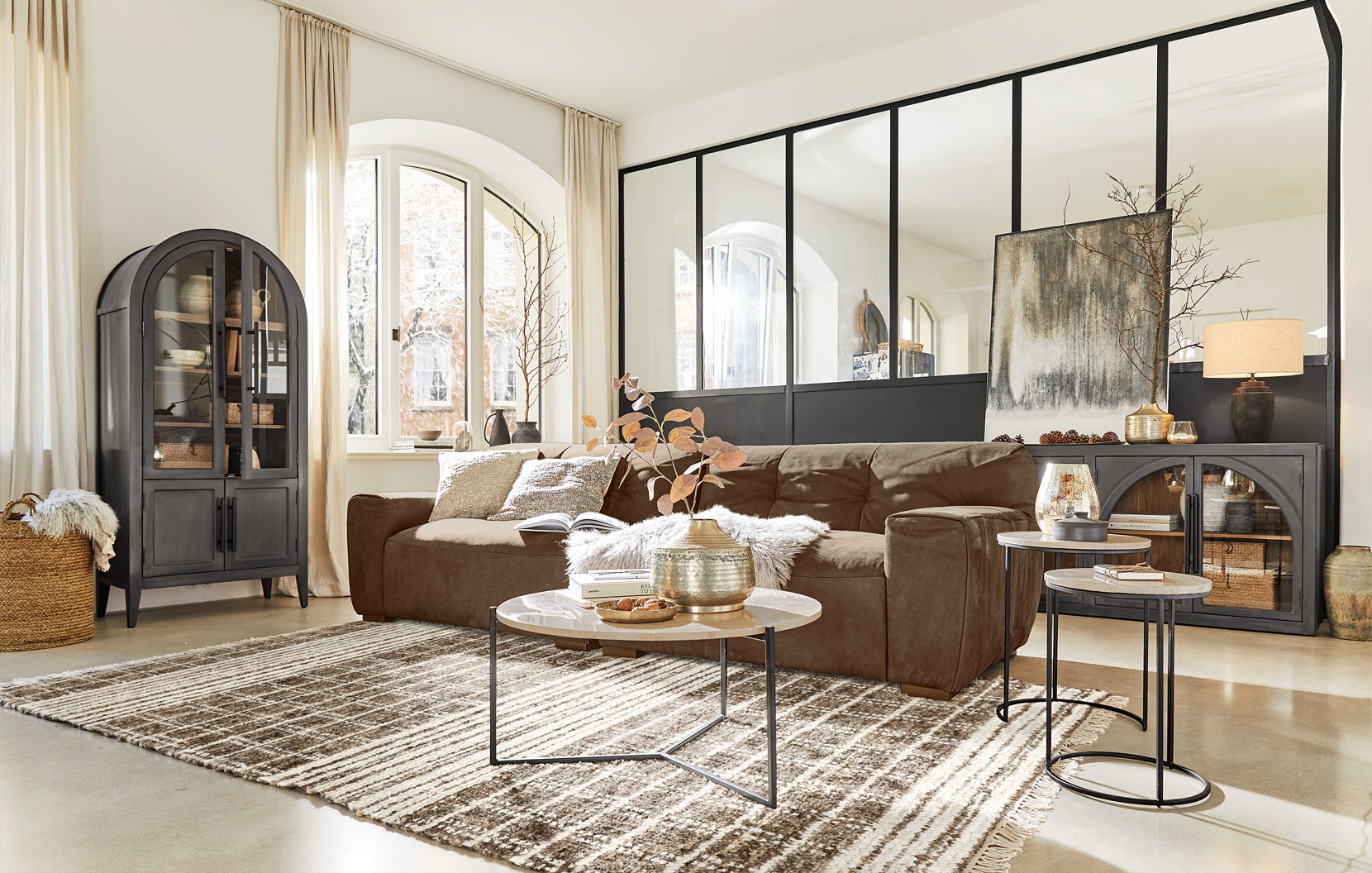 Gemütliches Wohnzimmer mit braunem Sofa, Metall-Beistelltischen und schwarzem Industrie-Glasvitrinenschrank