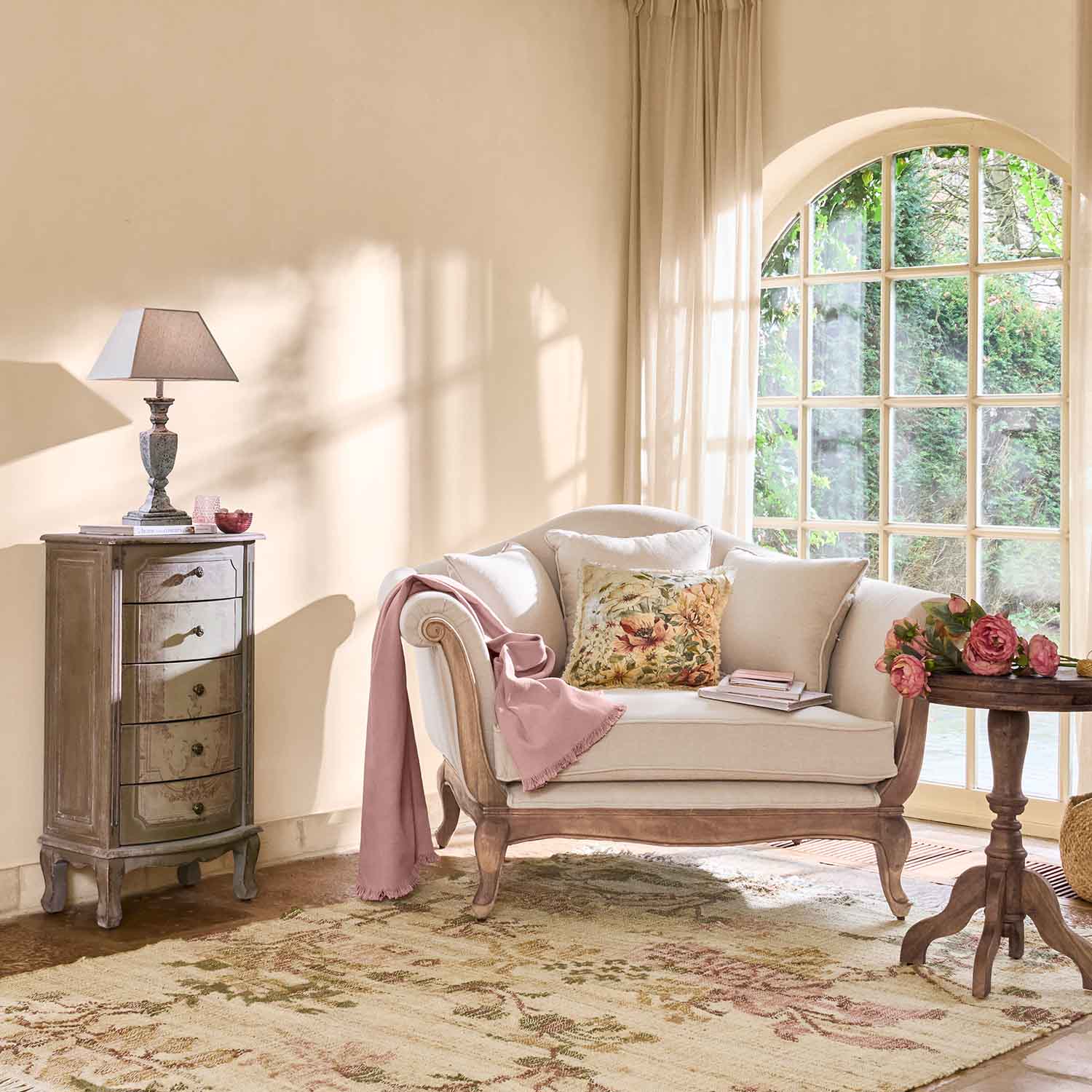 Cremefarbene Sofa mit Holzrahmen, Blumenmuster-Kissen und rosa Decke in hellem Wohnzimmer mit großem Fenster