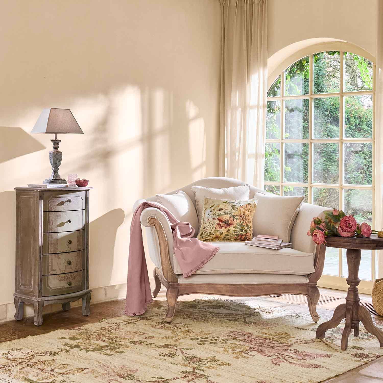 Cremefarbene Sofa mit Holzrahmen, Blumenmuster-Kissen und rosa Decke in hellem Wohnzimmer mit großem Fenster