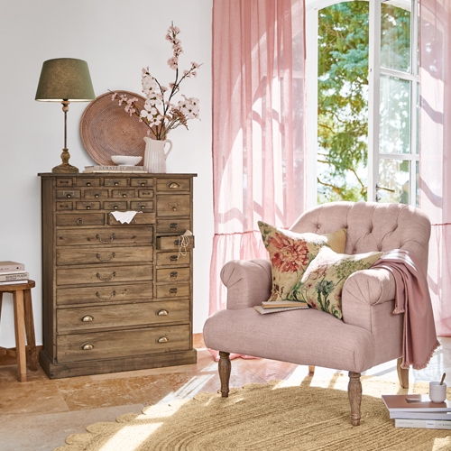 Gemütlicher rosa Sessel mit Blumen-Kissen neben einer Holzkommode in hellem Wohnzimmer mit rosa Vorhängen