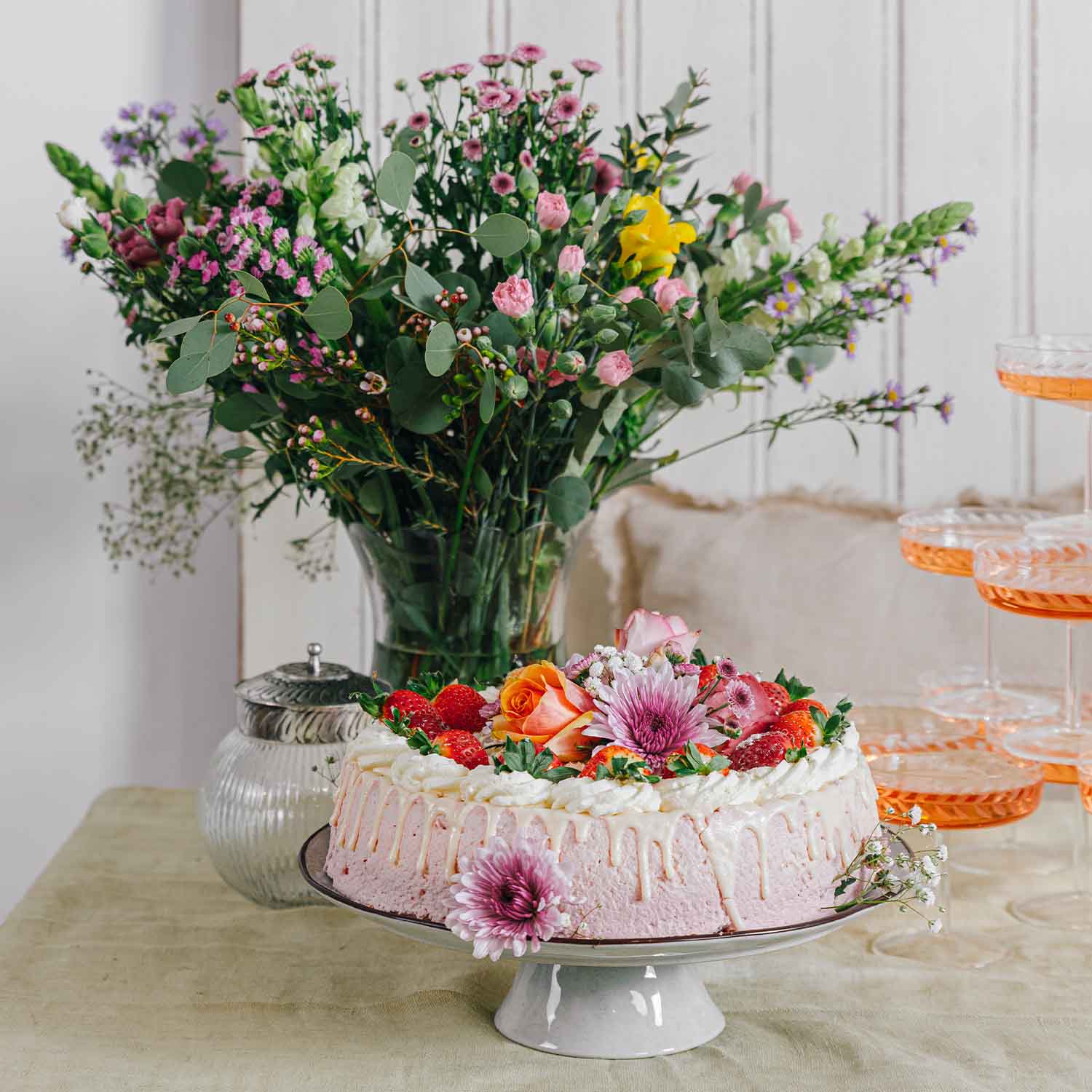 Rosafarbener Sahnetorte mit frischen Erdbeeren und Blumen auf weißem Tortenständer vor Blumenstrauß