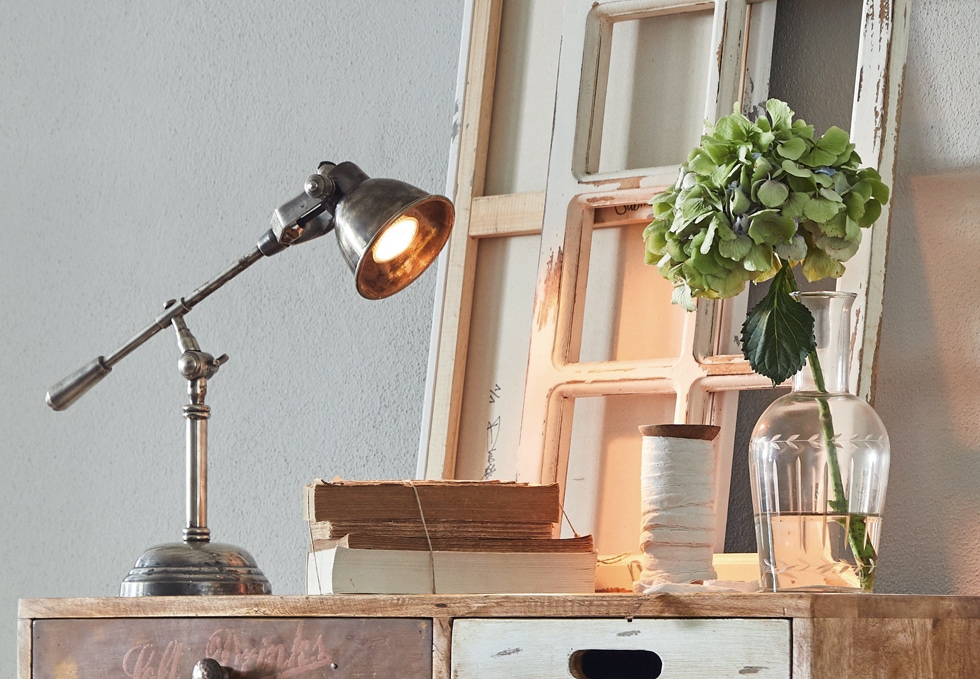 Industrielle Metall-Schreibtischlampe auf Holzkommode mit Büchern und Vase mit grünem Blumenstrauß