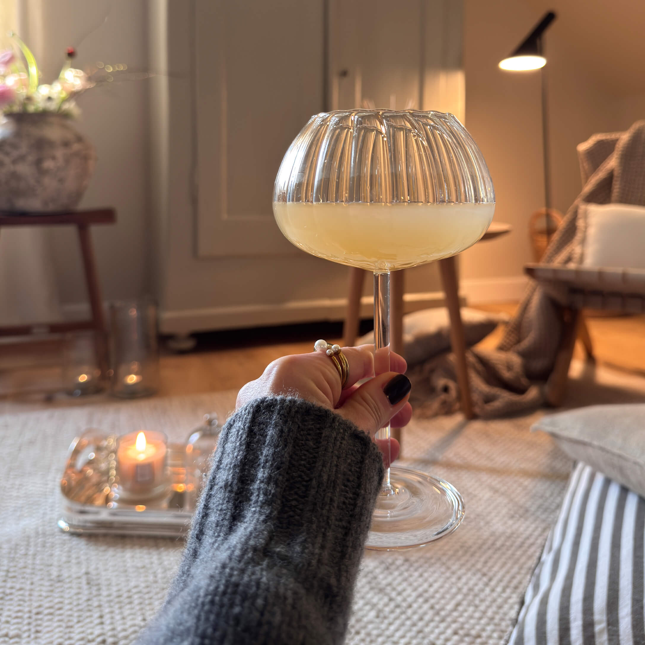 Hand hält ein Glas mit gelbem Getränk in gemütlichem Raum mit weicher Beleuchtung und Dekorationen