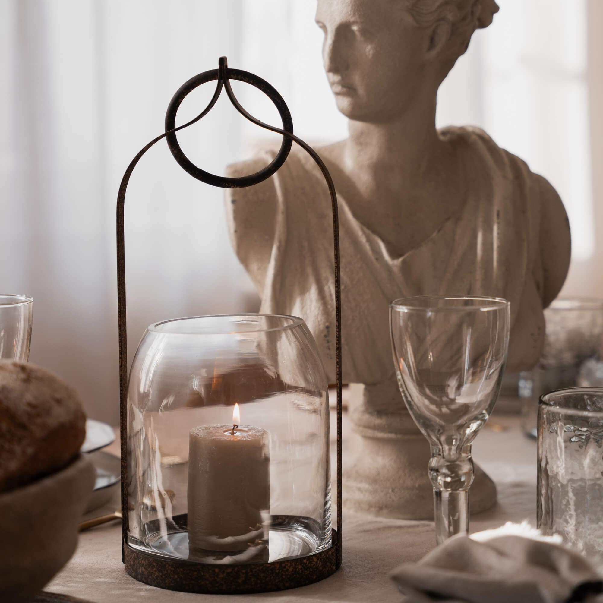 Glaslaterne mit rostiger Metallfassung und brennender Kerze auf rustikalem Tisch mit Skulptur