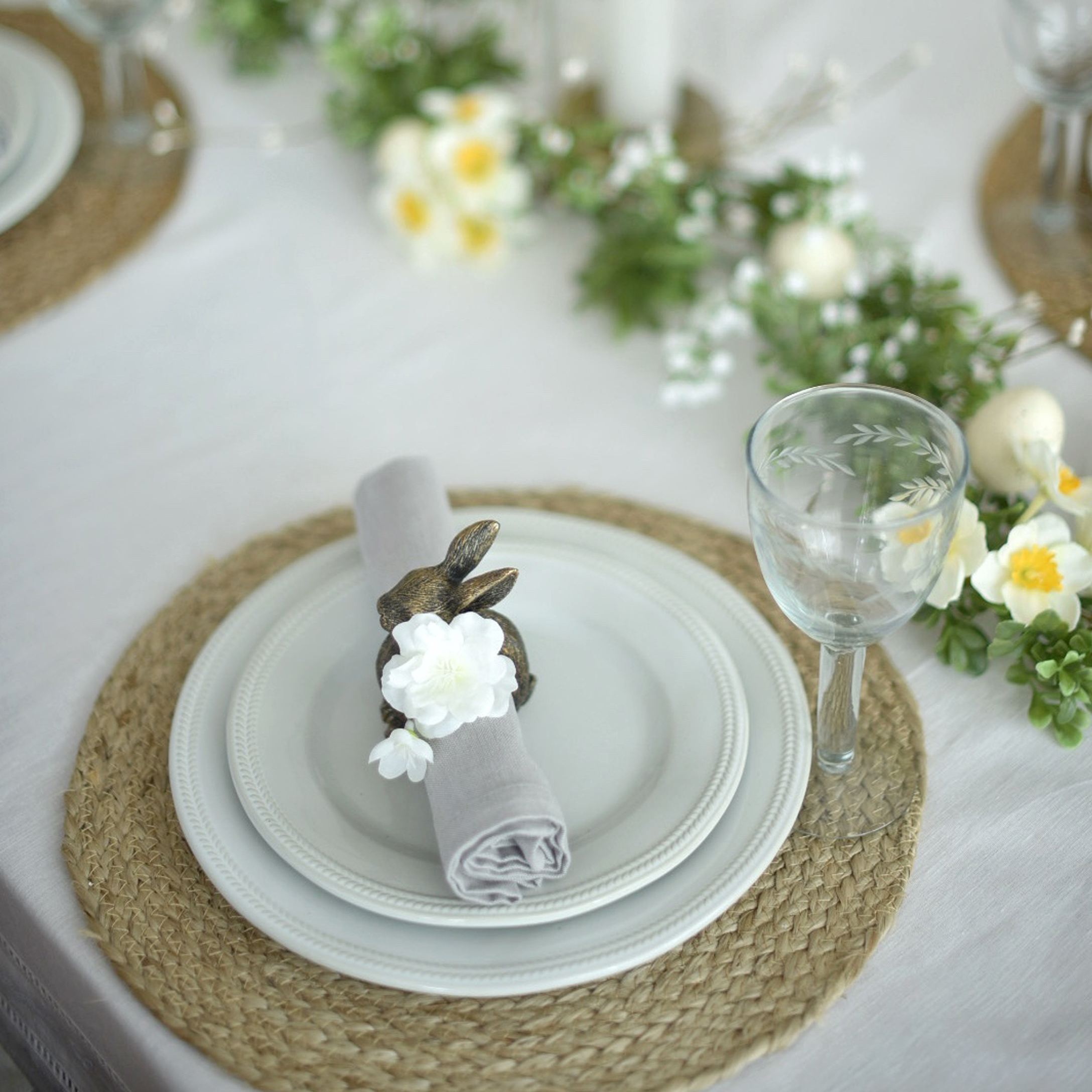Weißes Geschirr auf geflochtenem Platzset mit Hasen-Serviettenhalter und zarter Blumen-Dekoration