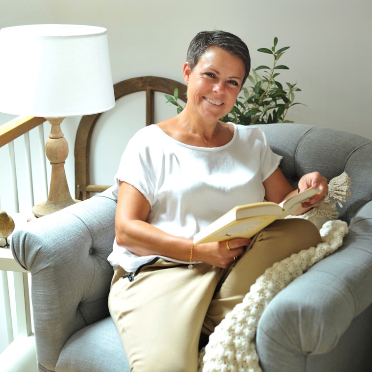 Frau in weißem Oberteil und beiger Hose liest Buch auf grauem Sofa in hellem Raum