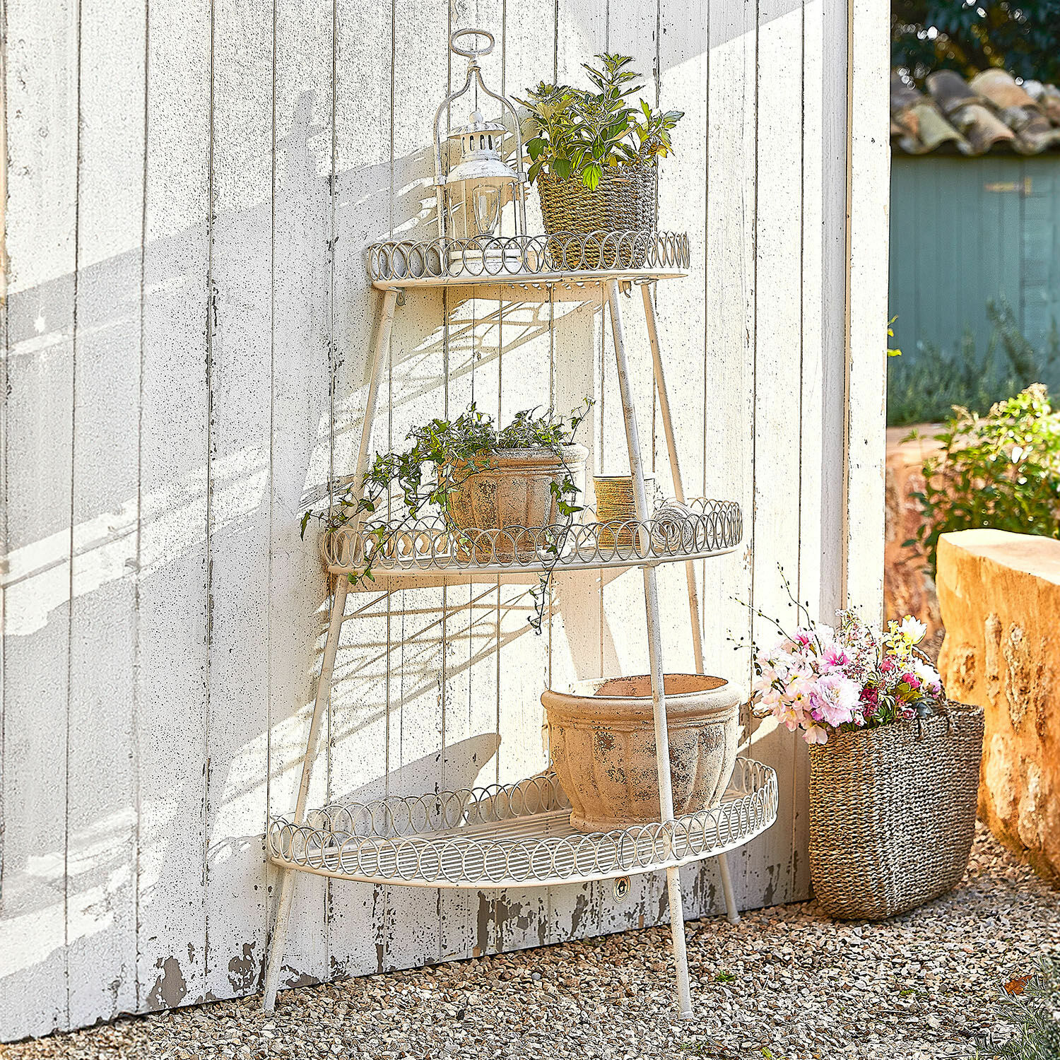 Etagere Lorivane Dreistufiges cremefarbenes Metallregal im Garten mit Blumentöpfen vor weißer Wand