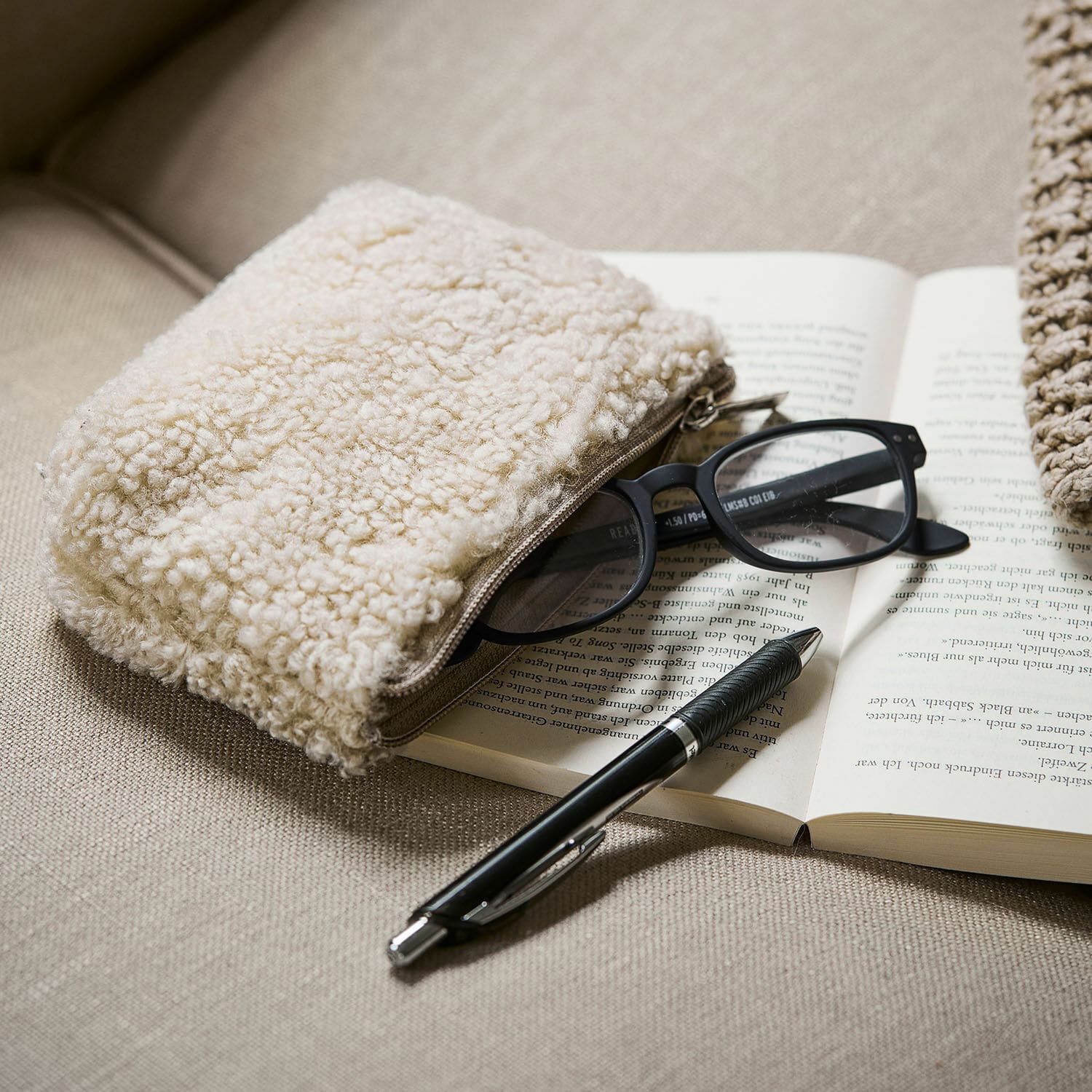 Täschchen Bathilde Beige Kosmetiktasche aus Teddy-Material auf Sofa mit Brille und schwarzem Kugelschreiber