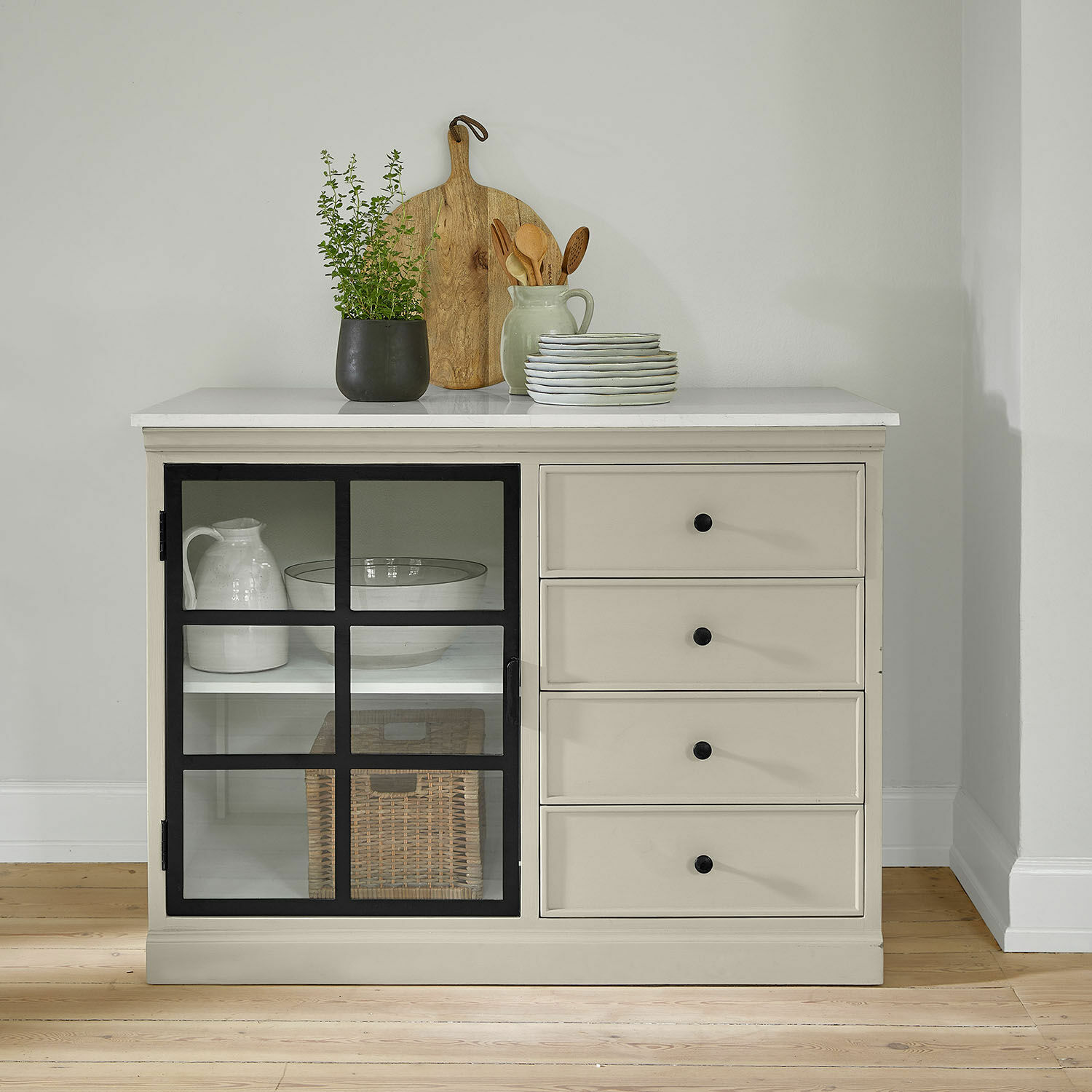 Schrank Orlévine Beige Kommode mit schwarzen Griffen und Glastür, dekoriert mit Holzbrett und grüner Pflanze