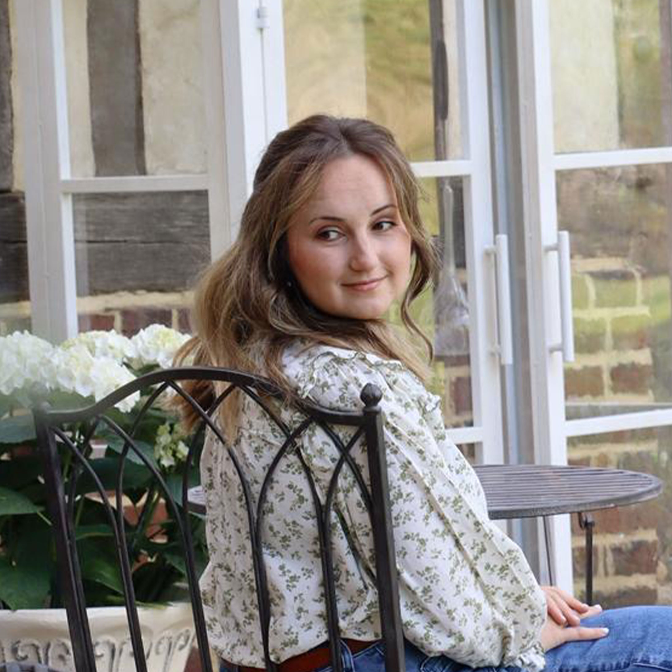 Frau in geblümter Bluse sitzt auf Metallstuhl in hellem Garten mit weißen Blumen