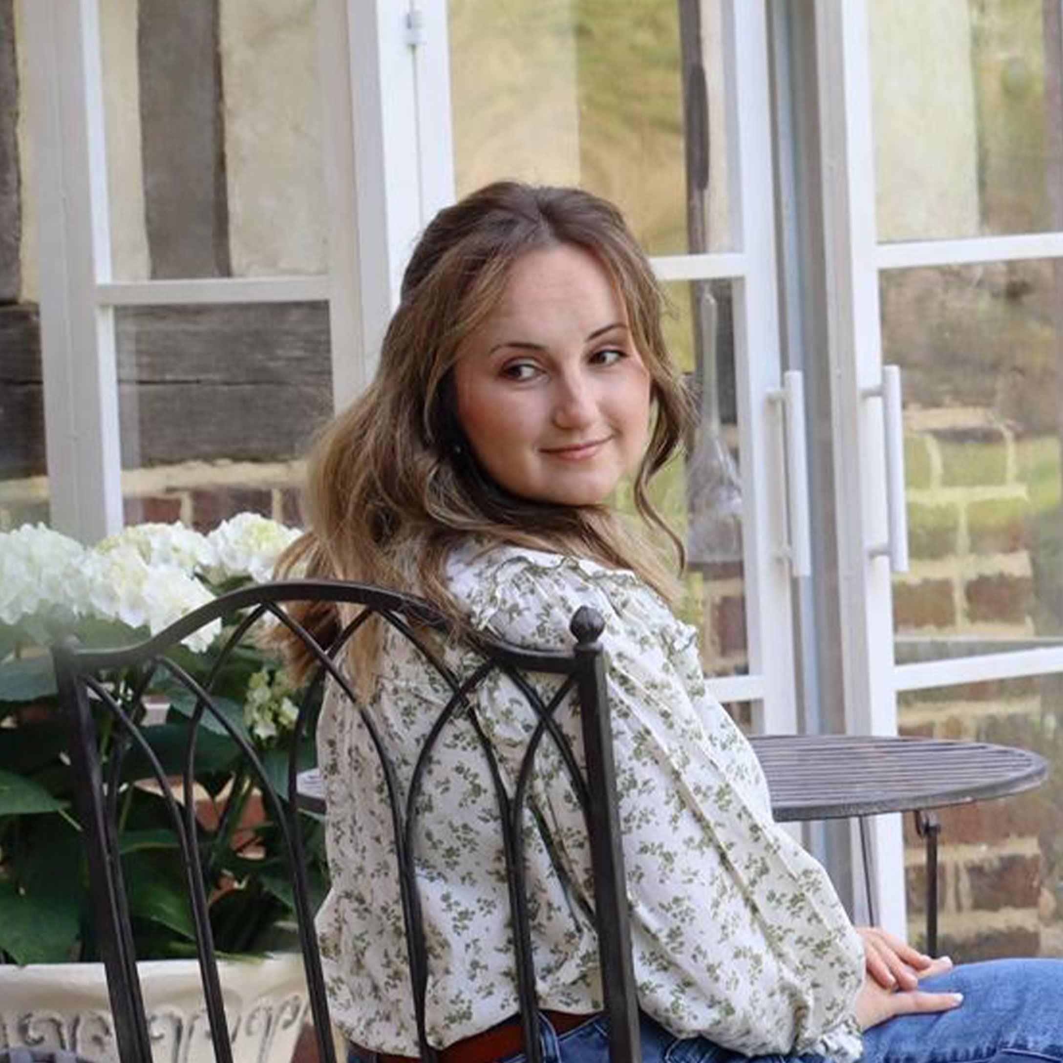 Frau in geblümter Bluse sitzt auf Metallstuhl in hellem Garten mit weißen Blumen