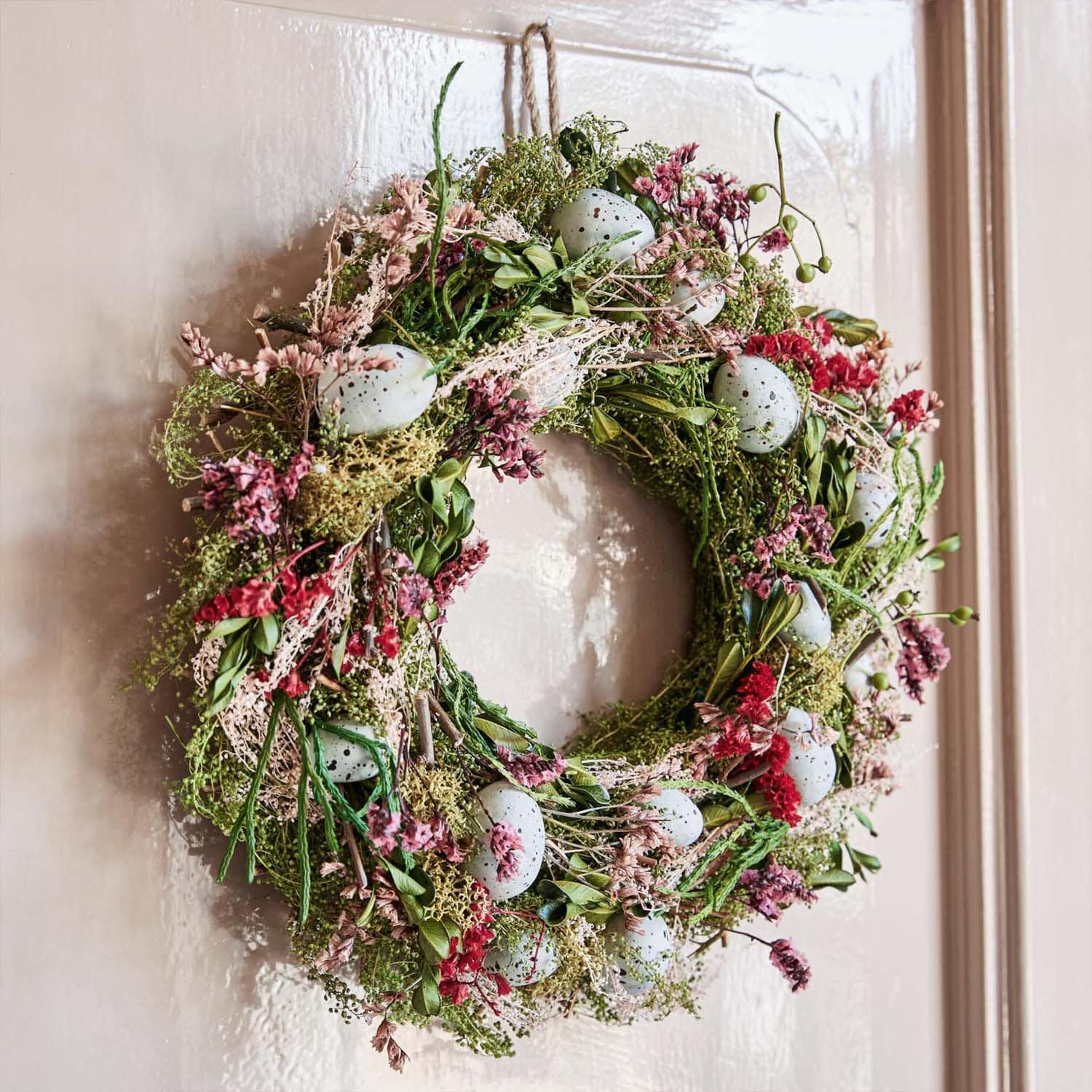 Dekorativer Kranz aus grünen Zweigen, rosa Blumen und blauen künstlichen Eiern an der Tür