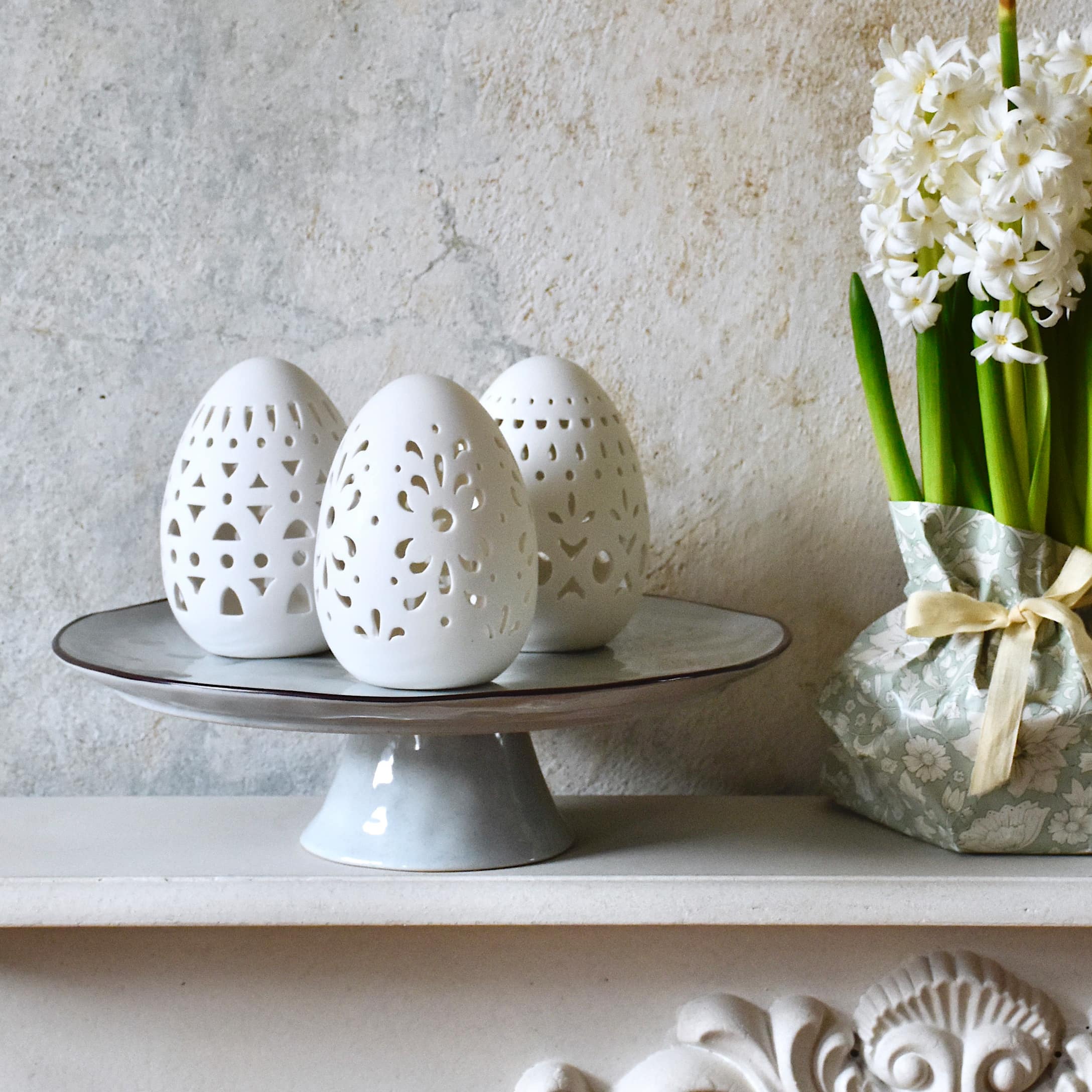 Weiße Keramik-Ostereier mit filigranen Mustern auf grauem Tortenständer, Frühlingsdeko in Pastellfarben