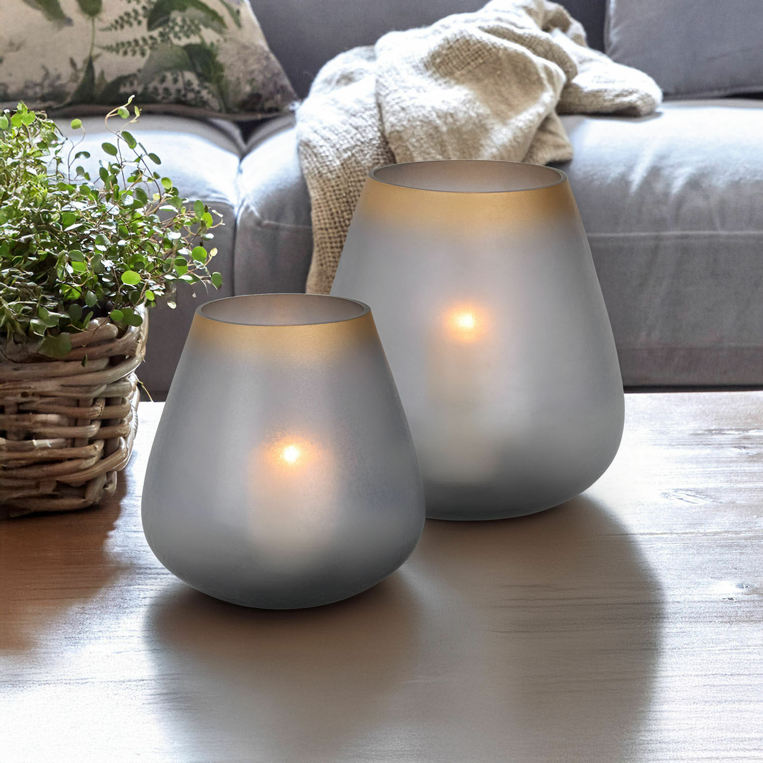 Zwei mattgraue Glas-Teelichthalter auf Holztisch im Wohnzimmer mit Pflanze und Sofa