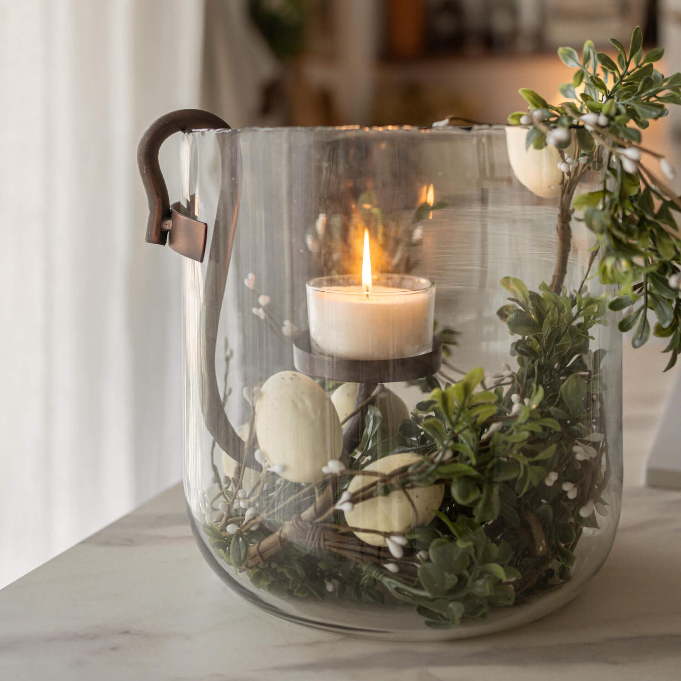 Glaslaterne mit Metallgriff, brennende weiße Kerze und grüne Blattdekoration mit weißen Beeren