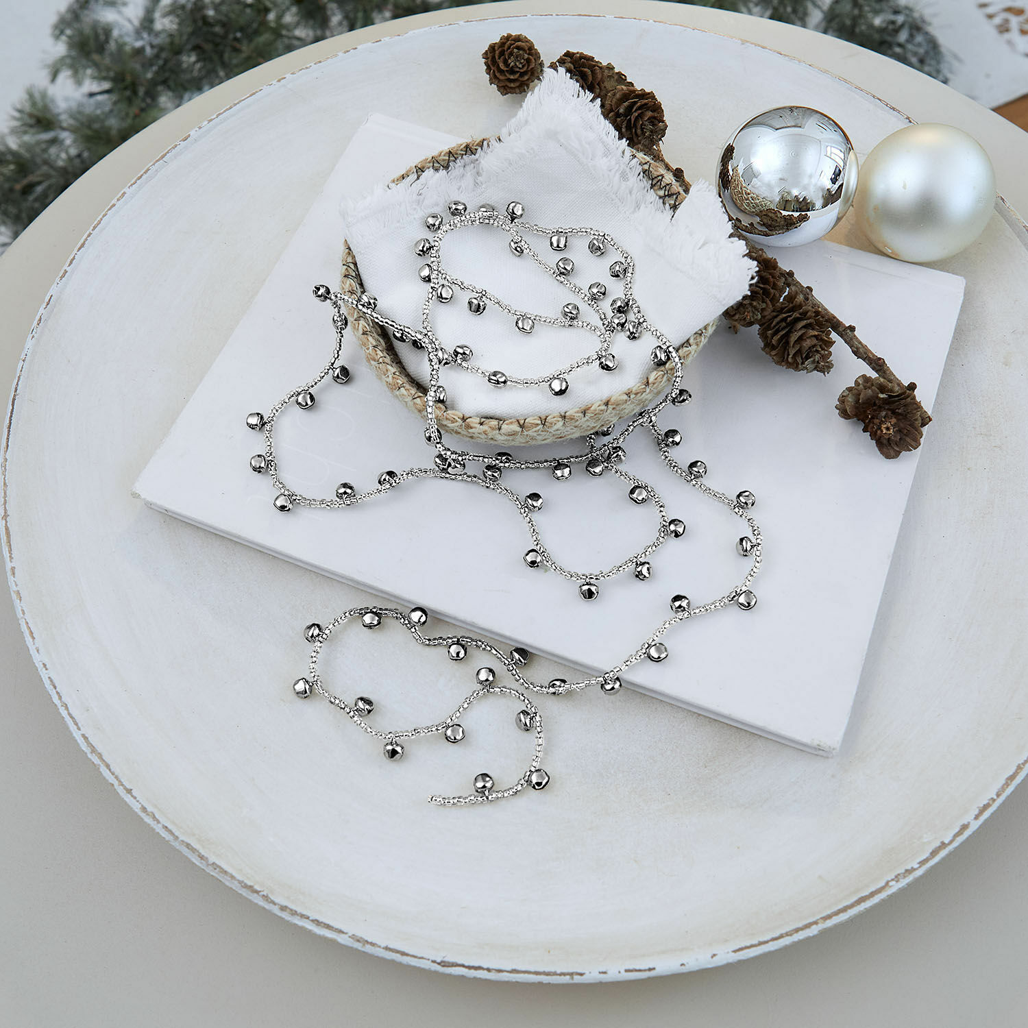 Silberne Kette mit Glöckchen auf weißem Hintergrund mit Weihnachtsdeko und Zapfen