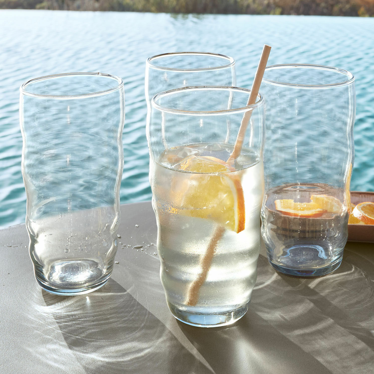 Glas 4er Set Tournavin Vier durchsichtige, wellenförmige Gläser auf Tisch am Wasser, ein Glas mit Zitrone und Strohhalm