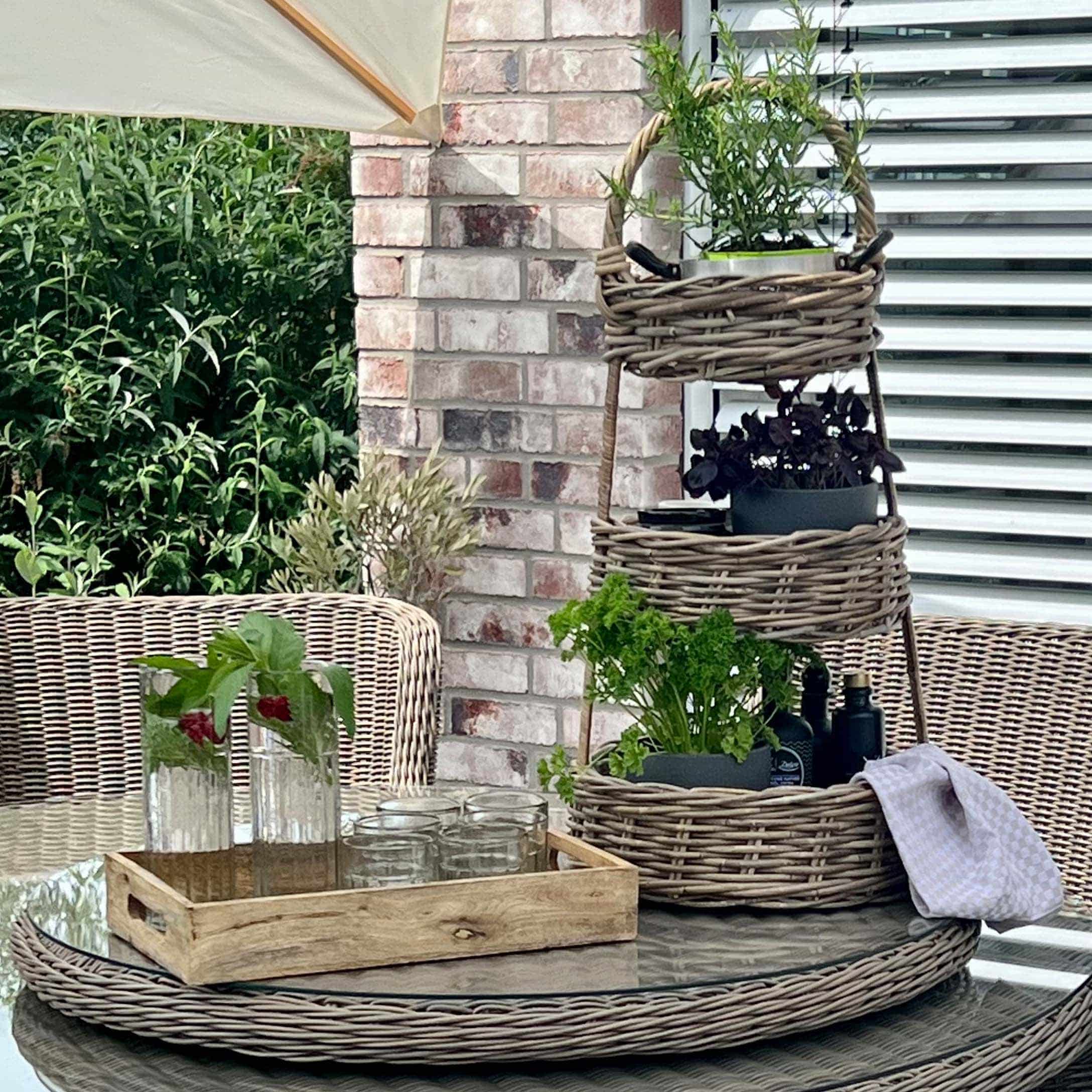 Dreistöckiges Rattanständer mit grünen Pflanzen auf Glastisch auf Terrasse mit Rattanmöbeln