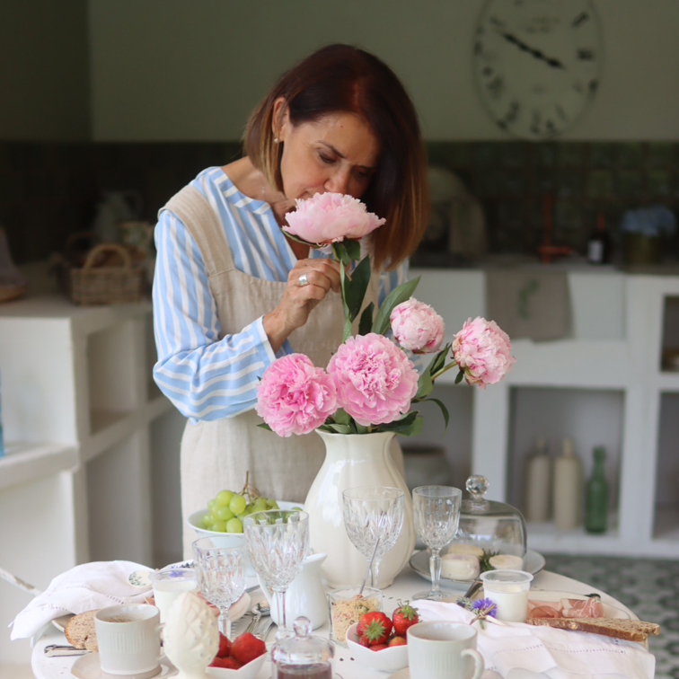Frau in Küche riecht an rosa Pfingstrosen in weißer Vase auf gedecktem Tisch mit Essen und Gläsern