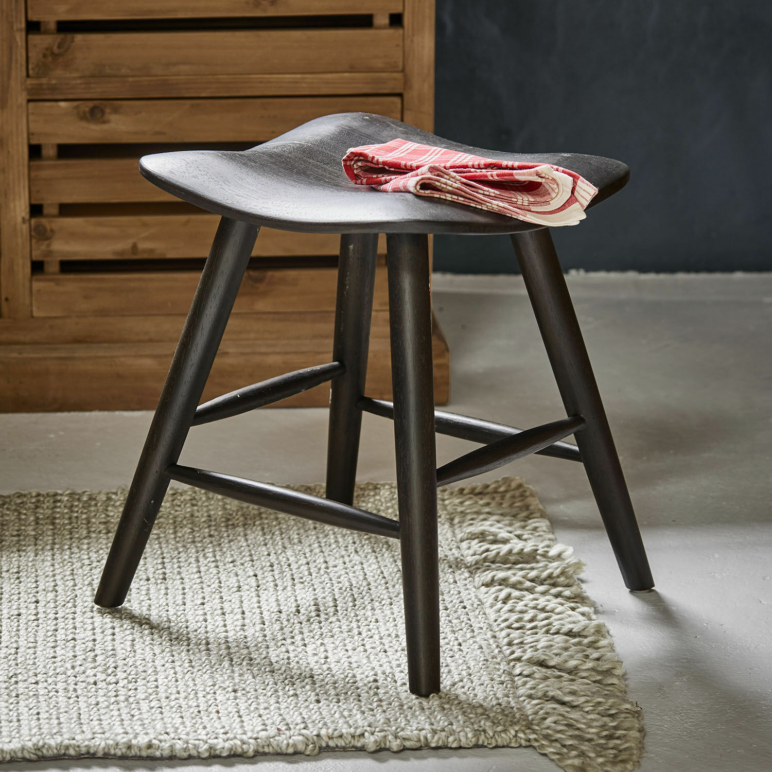 Schwarzer Holzschemel mit geschwungenem Sitz auf hellem Teppich, rote dekorative Serviette