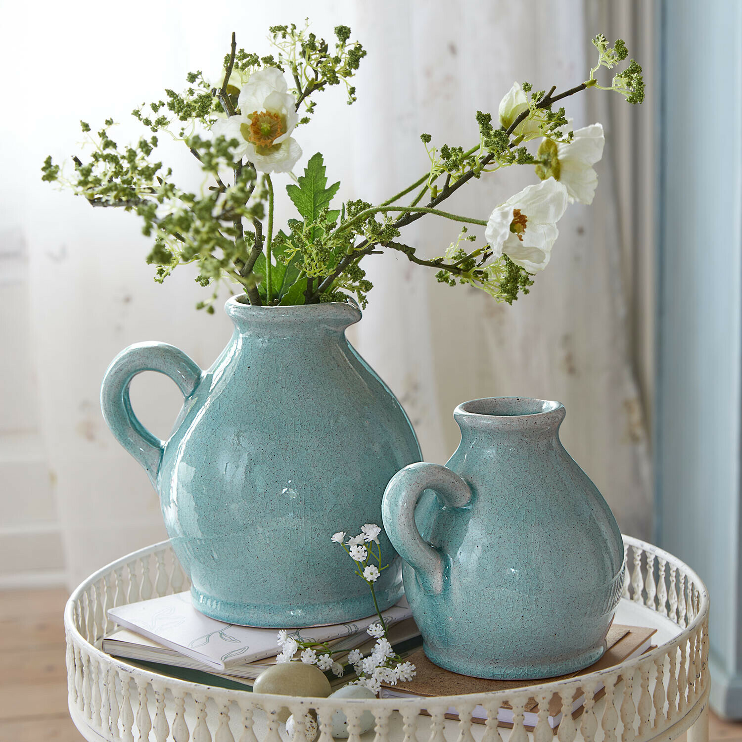 Zwei türkisfarbene Keramikkrüge mit Griffen auf weißem Metalltisch mit Blumen und Büchern
