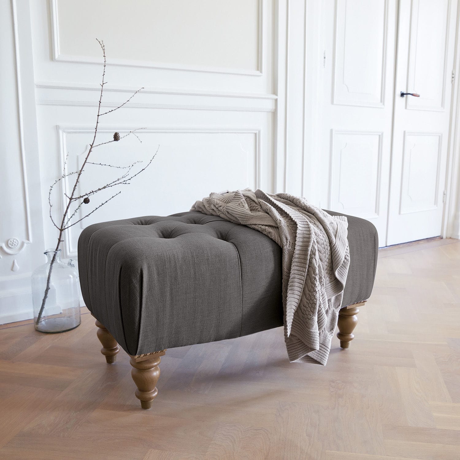 Hocker Durance Grau gepolsterter Hocker mit Holzbeinen und beiger Decke in hellem Raum
