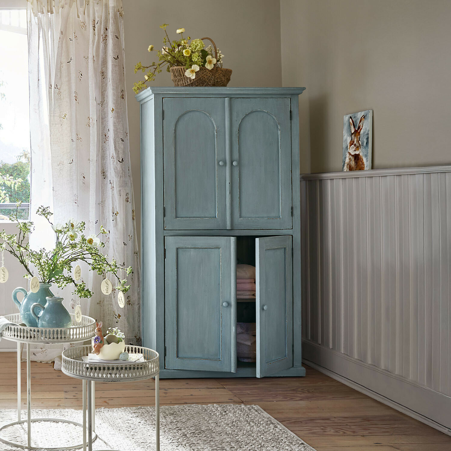 Blauer Holzschrank mit zwei Türen in gemütlichem Wohnzimmer mit Frühlingsdekoration und Blumen