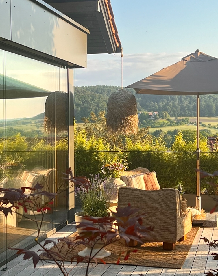 Gemütliche Terrasse mit beigem Sofa, Sonnenschirm und Pflanzen vor grünem Landschaftshintergrund, Gartenmöbel