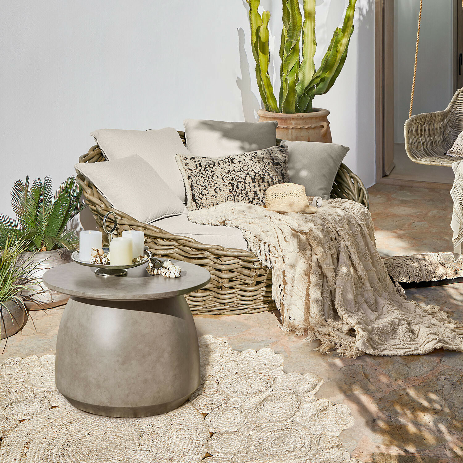 Rundes Rattansofa mit cremefarbenen Kissen und Decke auf Terrasse mit Beton-Couchtisch