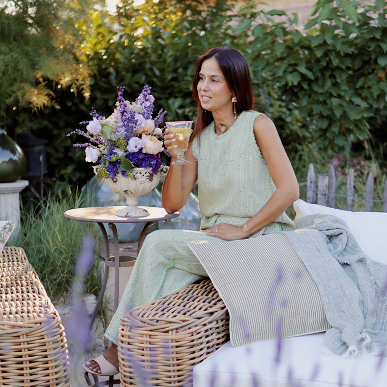 Frau im grünen Kleid sitzt auf Rattanmöbel im Garten mit Blumenstrauß und Weinglas