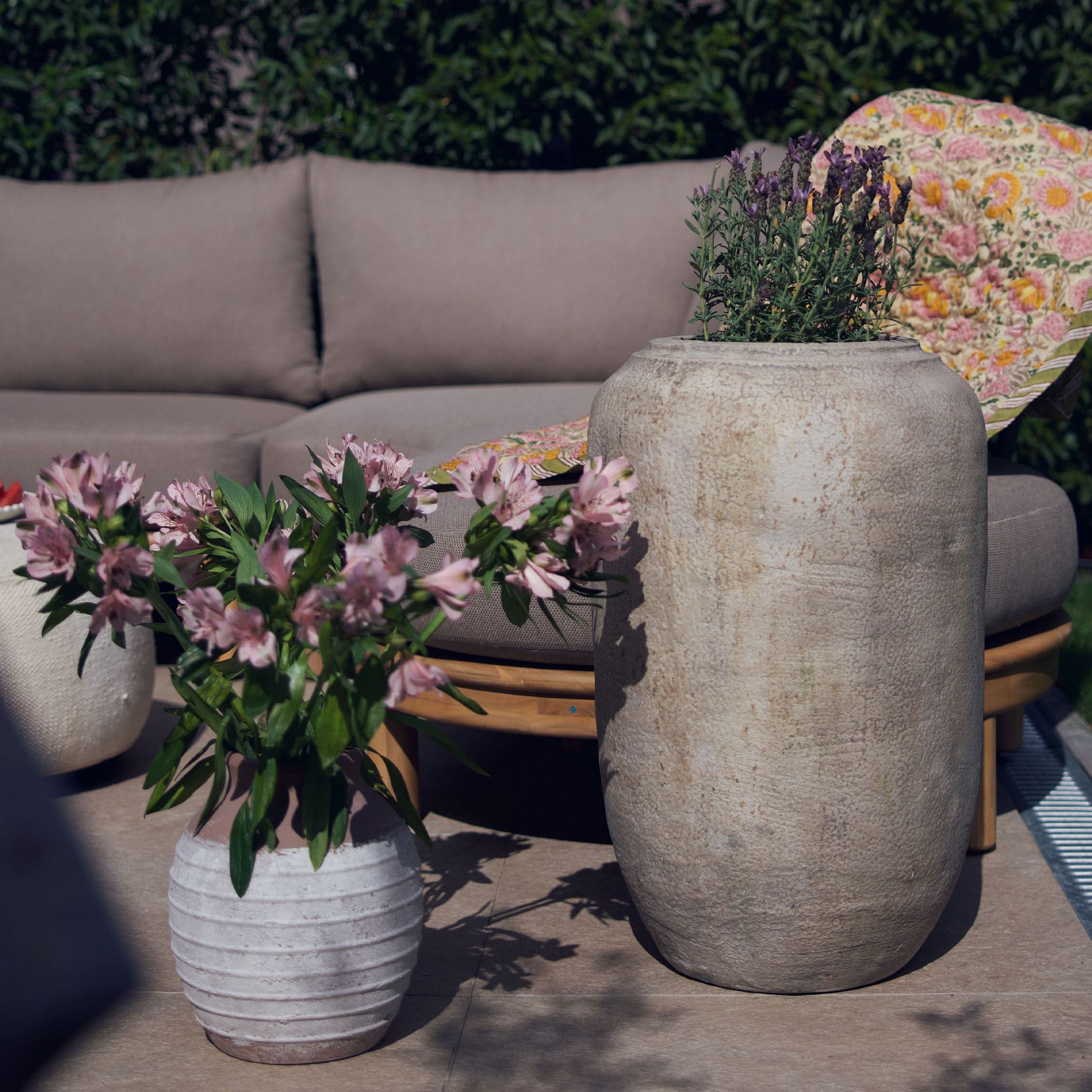 Großer und kleiner heller Steintopf mit Blumen auf Terrasse mit grauem Sofa und buntem Plaid