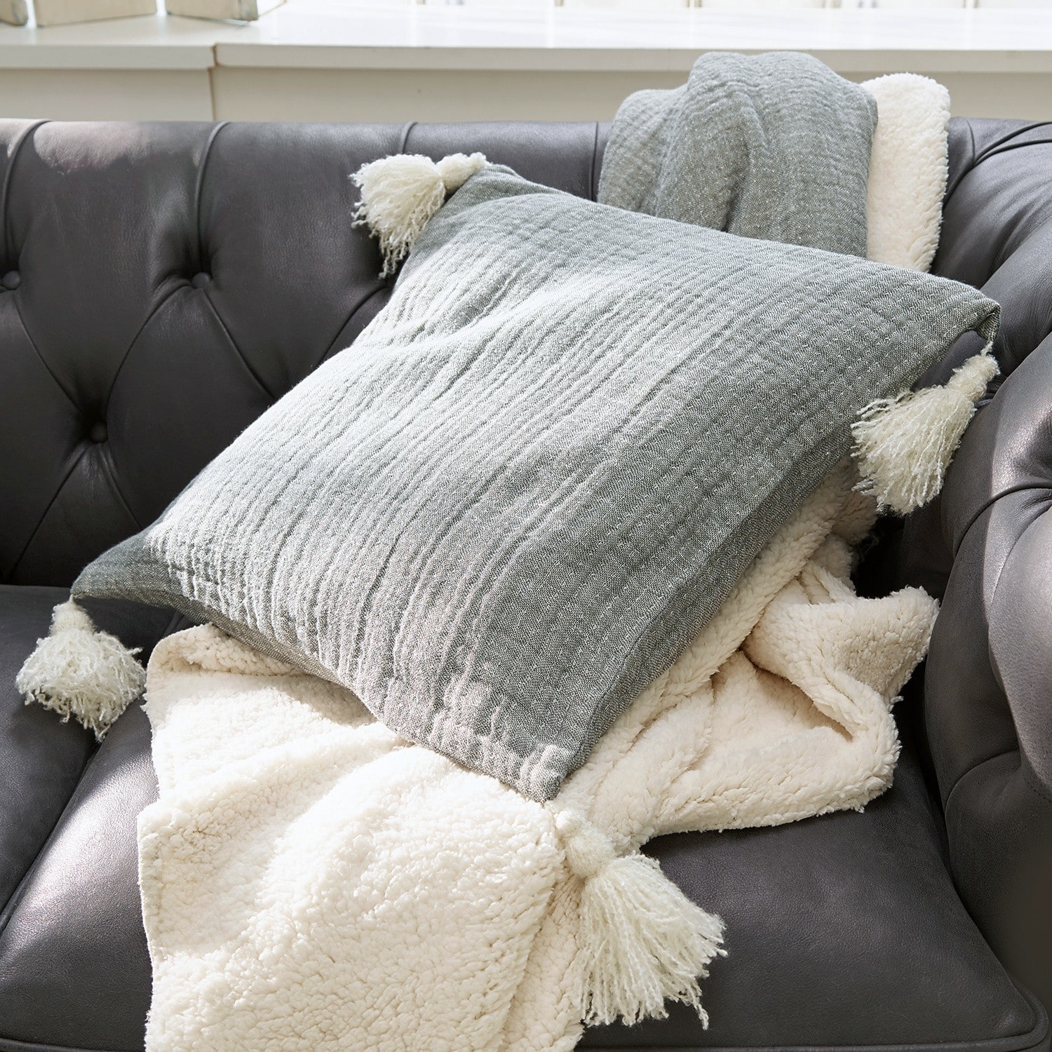 Kissen Malliévre Graues Kissen mit Quasten und cremefarbene weiche Decke auf dunklem Ledersofa im Wohnzimmer