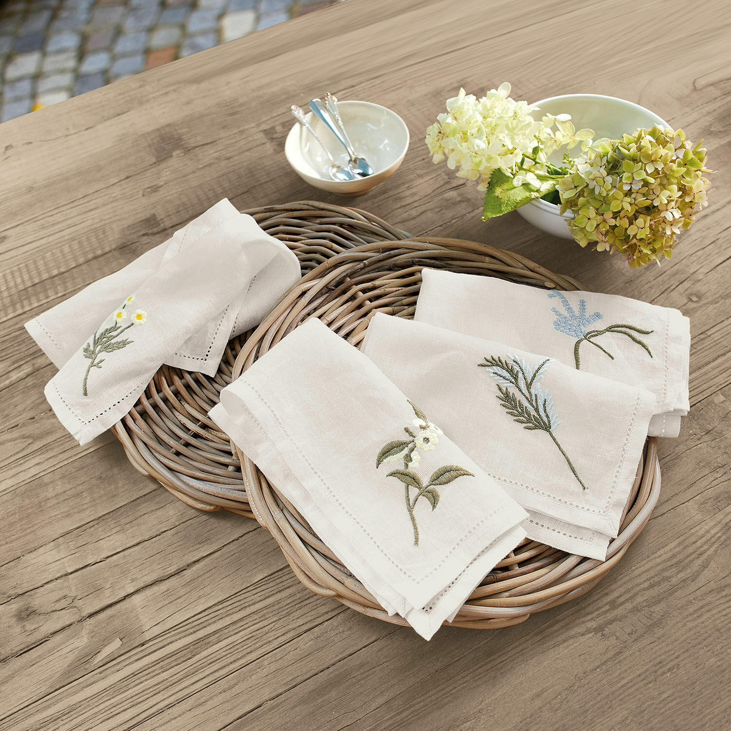 Serviette 4er Set Lentilly Leinenservietten mit pflanzlicher Stickerei in Naturfarbe auf einem Korb aus Weide auf Holztisch