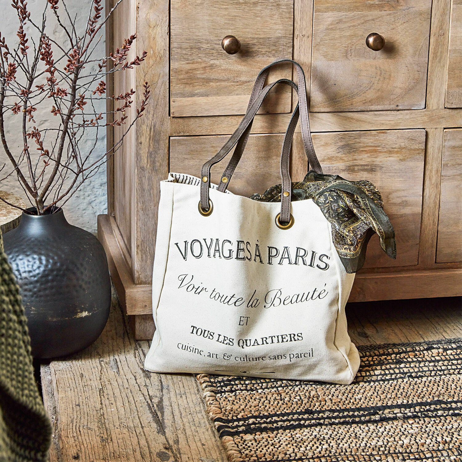 Tasche Voyage Beige Baumwolltasche mit schwarzem Aufdruck und braunen Griffen auf Holzboden