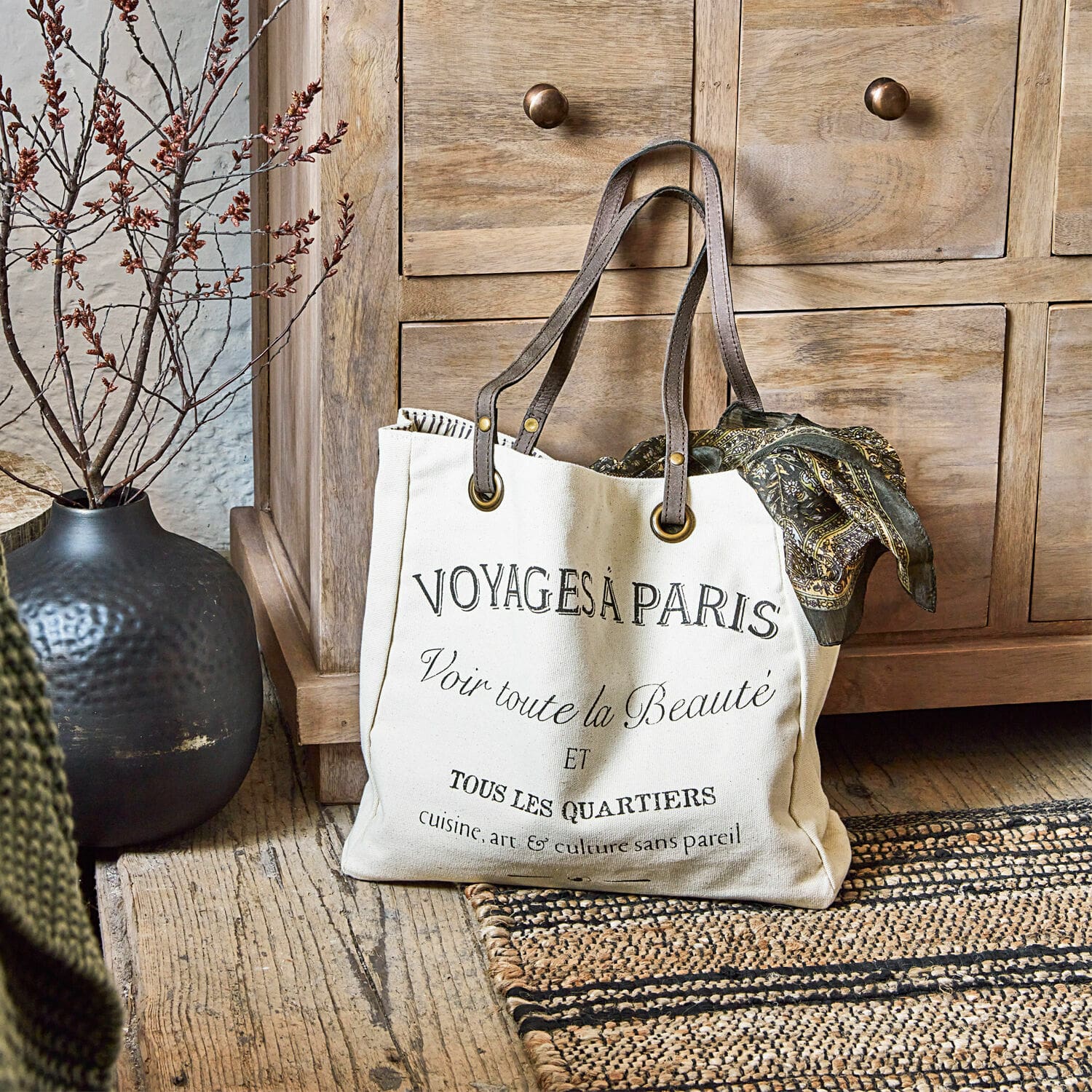 Tasche Voyage Beige Baumwolltasche mit schwarzem Aufdruck und braunen Griffen auf Holzboden