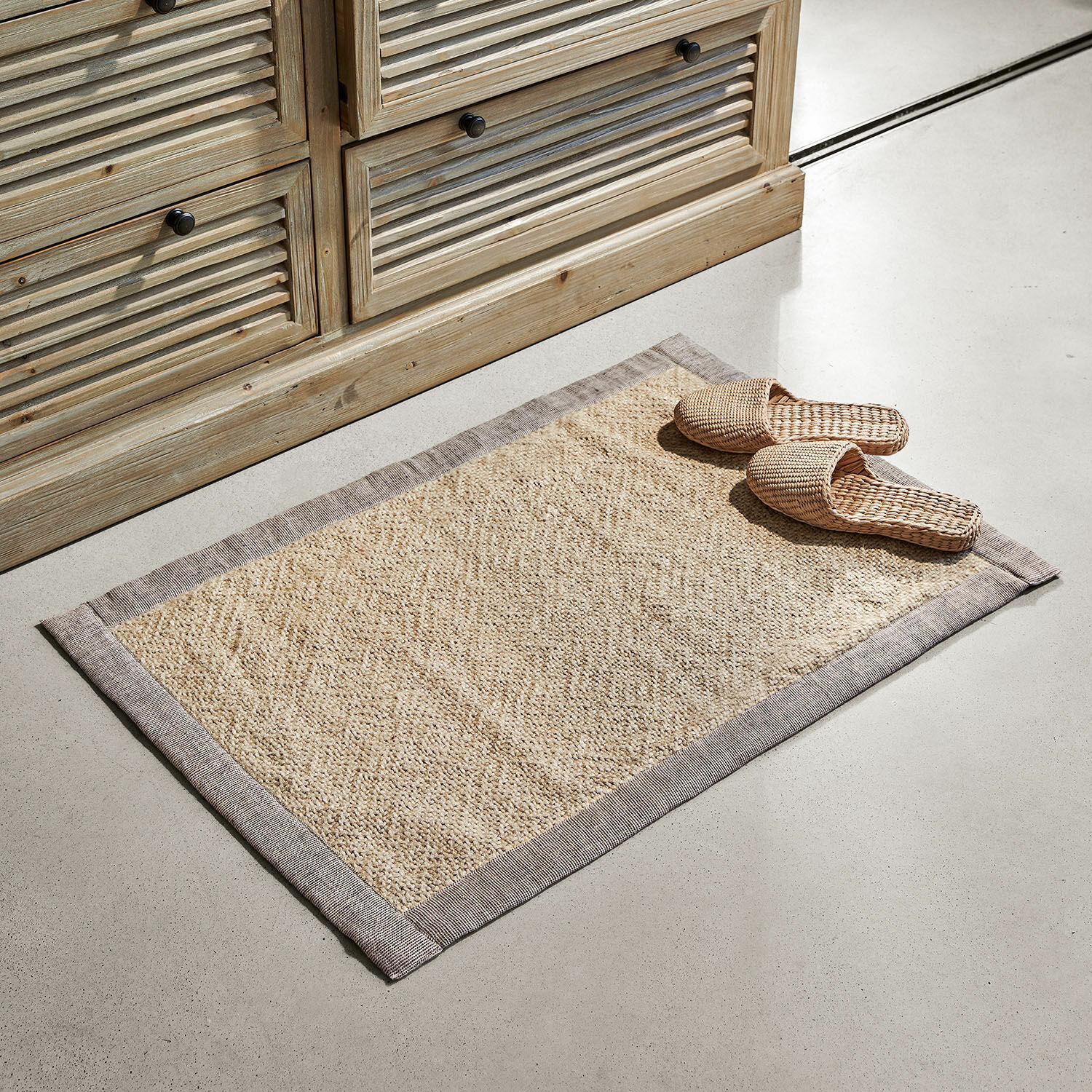 Badematte Wilder Beiger Teppich aus Naturmaterial mit grauem Rand auf hellem Boden vor Holzkommode