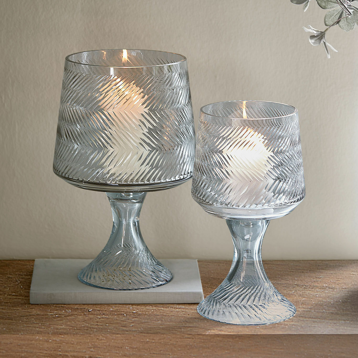 Zwei transparente Glaswindlichter mit Reliefmuster und weißen Kerzen auf Holztisch