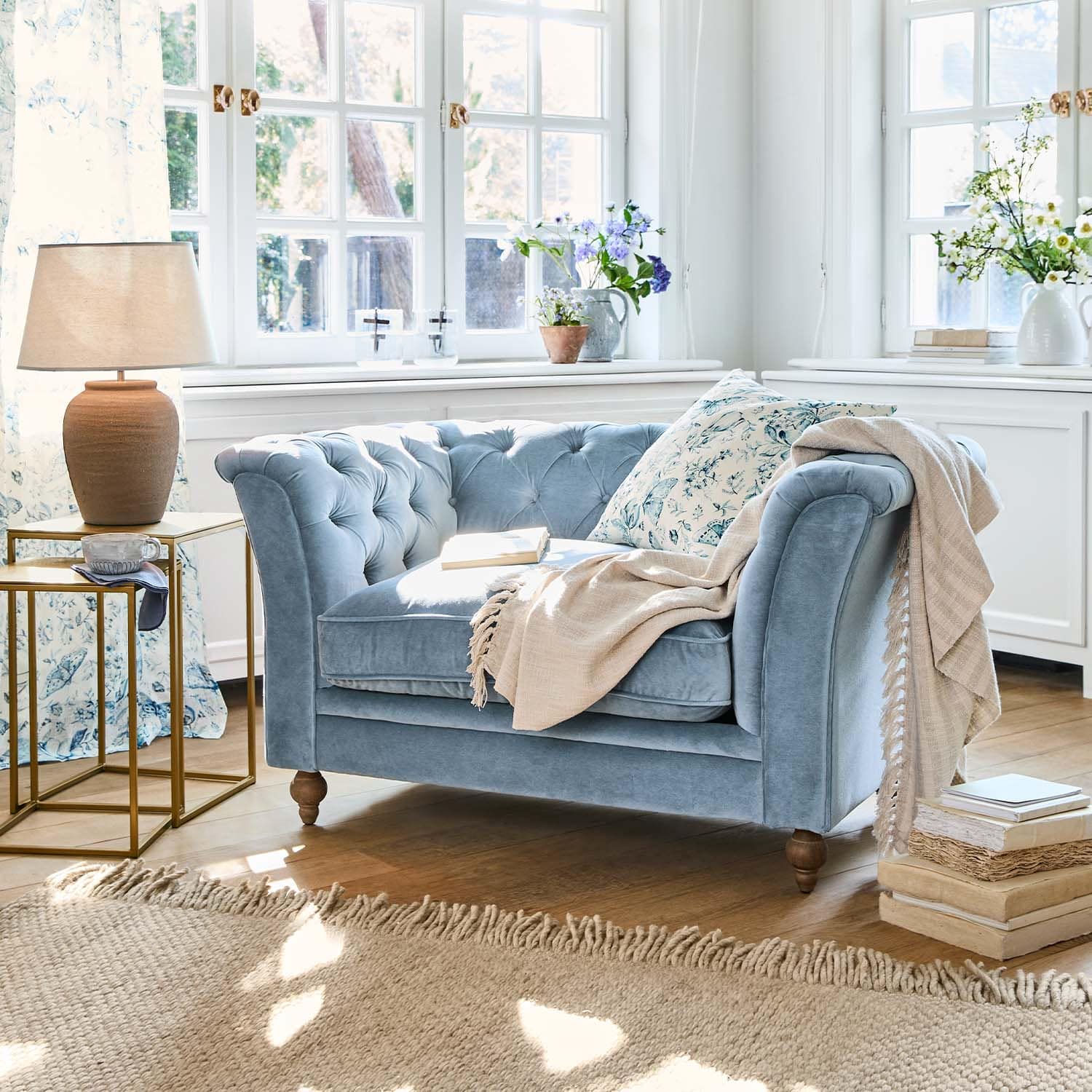 Zweisitziges blaues Samtsofa mit floralem Kissen und beiger Decke in hellem Wohnzimmer mit Holzboden