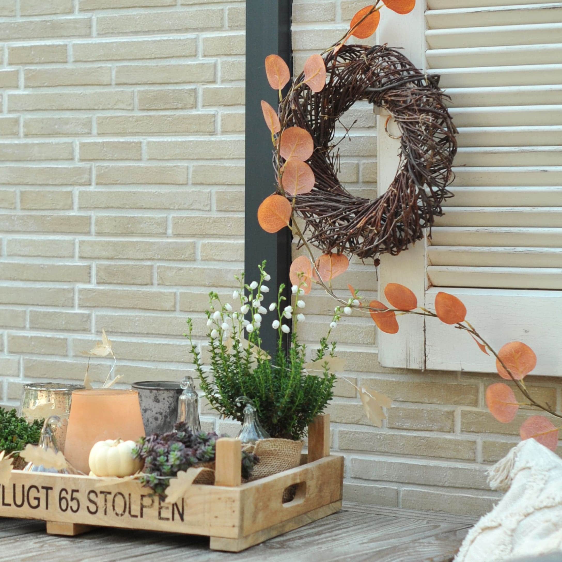 Holztablett mit herbstlicher Pflanzen- und Laternen-Dekoration vor Ziegelwand