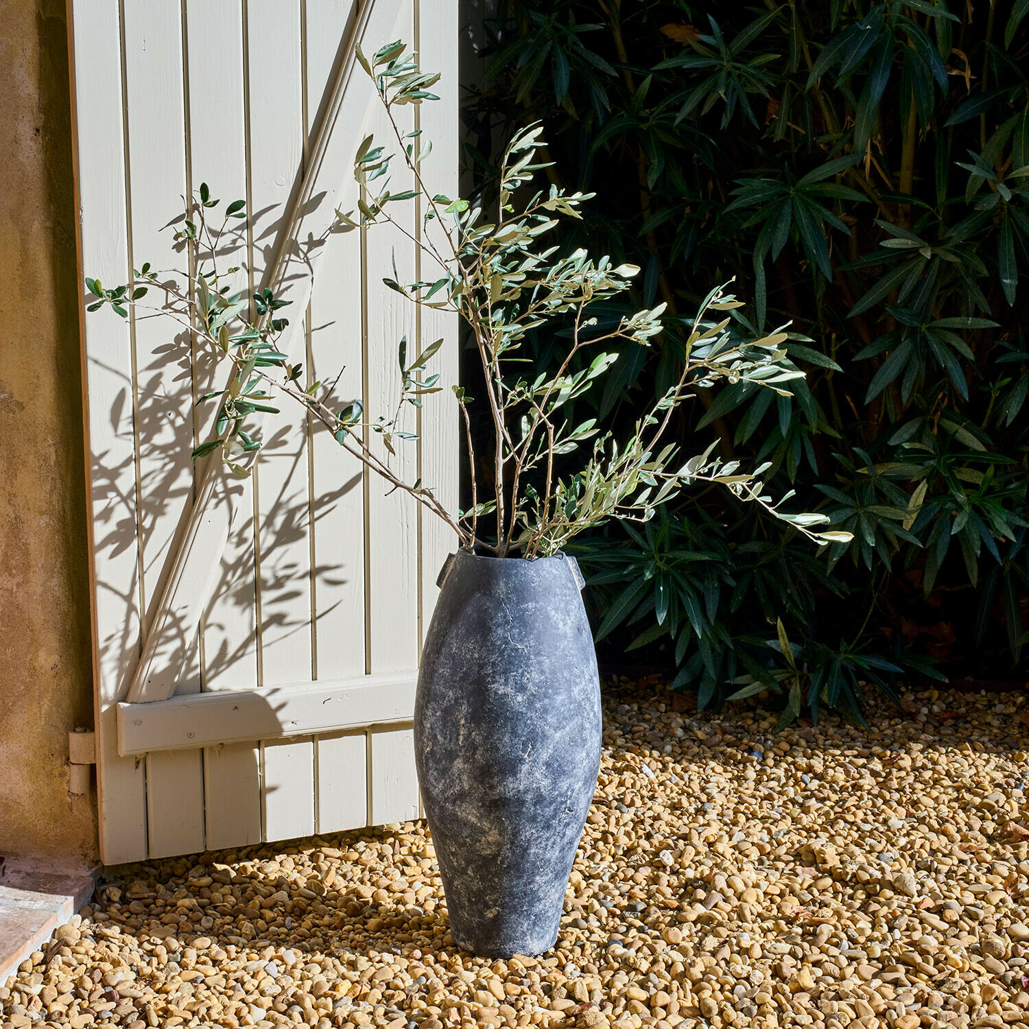 Hohe graue Steinvase mit Olivenzweigen auf Kies vor weißer Tür