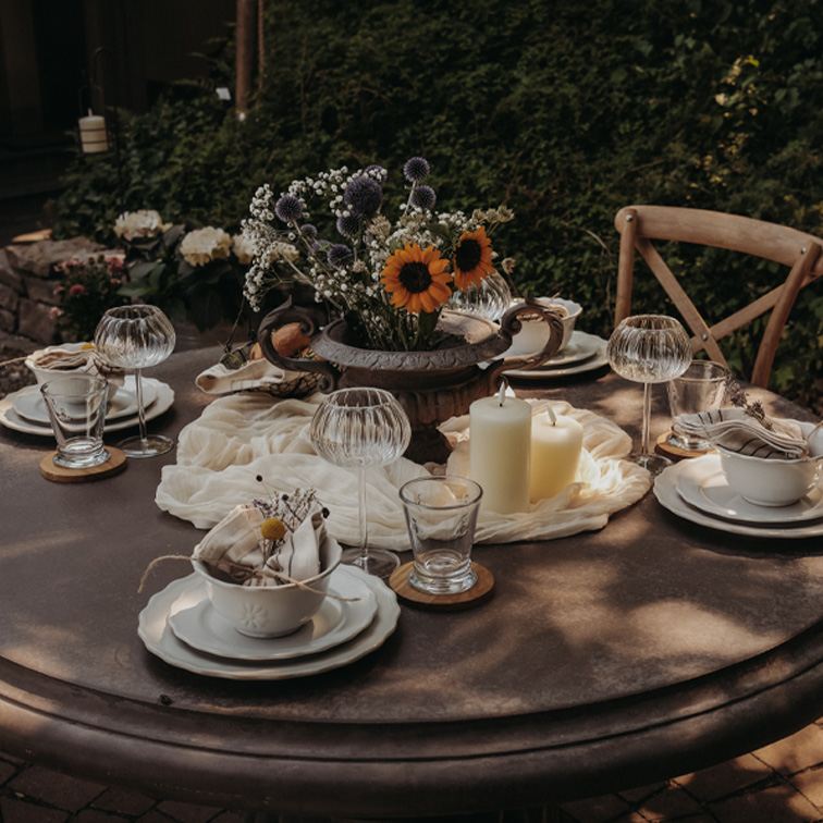 Runder Gartentisch mit Kerzen und Blumenarrangement in Metallvase für eine festliche Tafel.