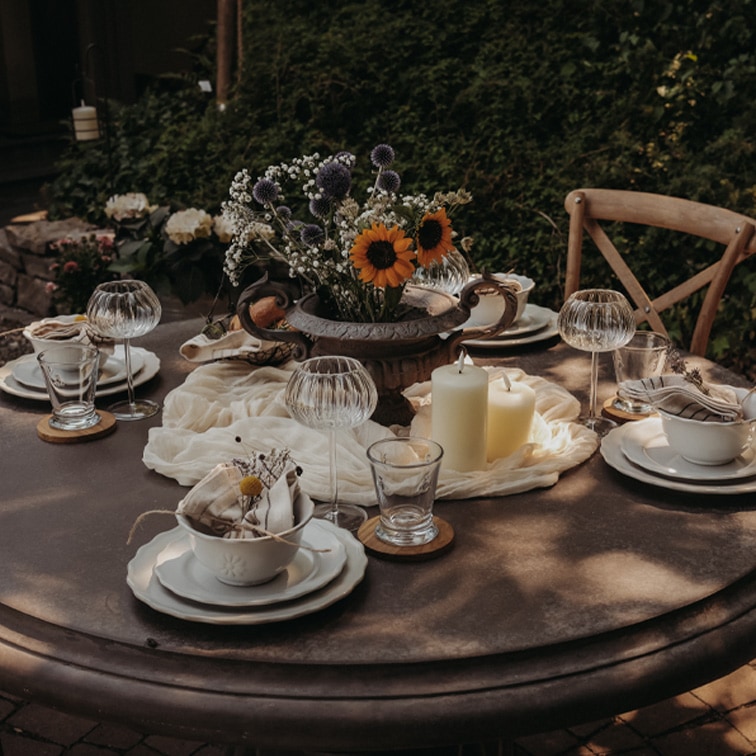 Runder Gartentisch mit Kerzen und Blumenarrangement in Metallvase für eine festliche Tafel.